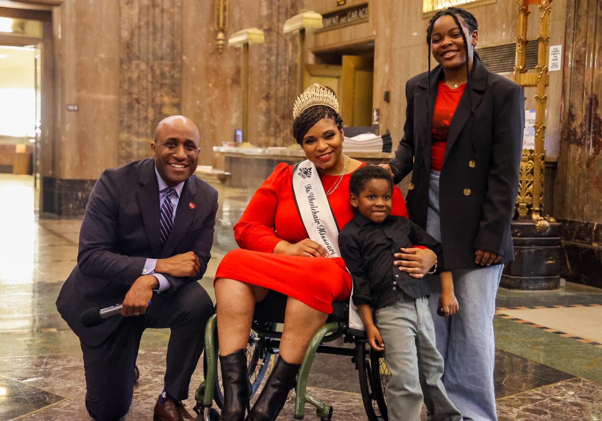Four people pose indoors, one sitting in a wheelchair, smiling, with a woman wearing a sash, and two standing women.