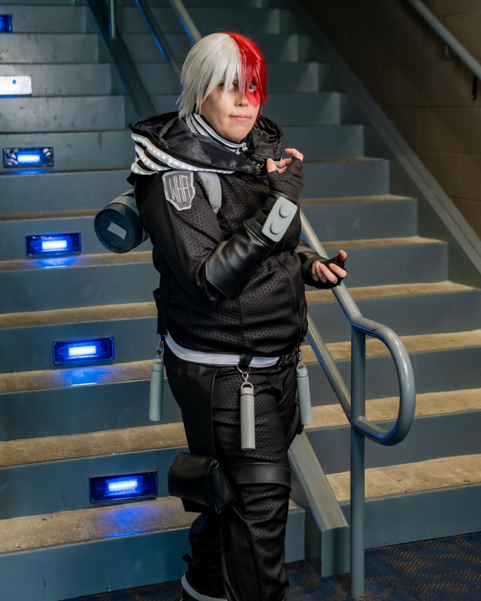 Person dressed as a character with white and red hair, black outfit, standing on stairs with blue lights, holding a prop weapon.