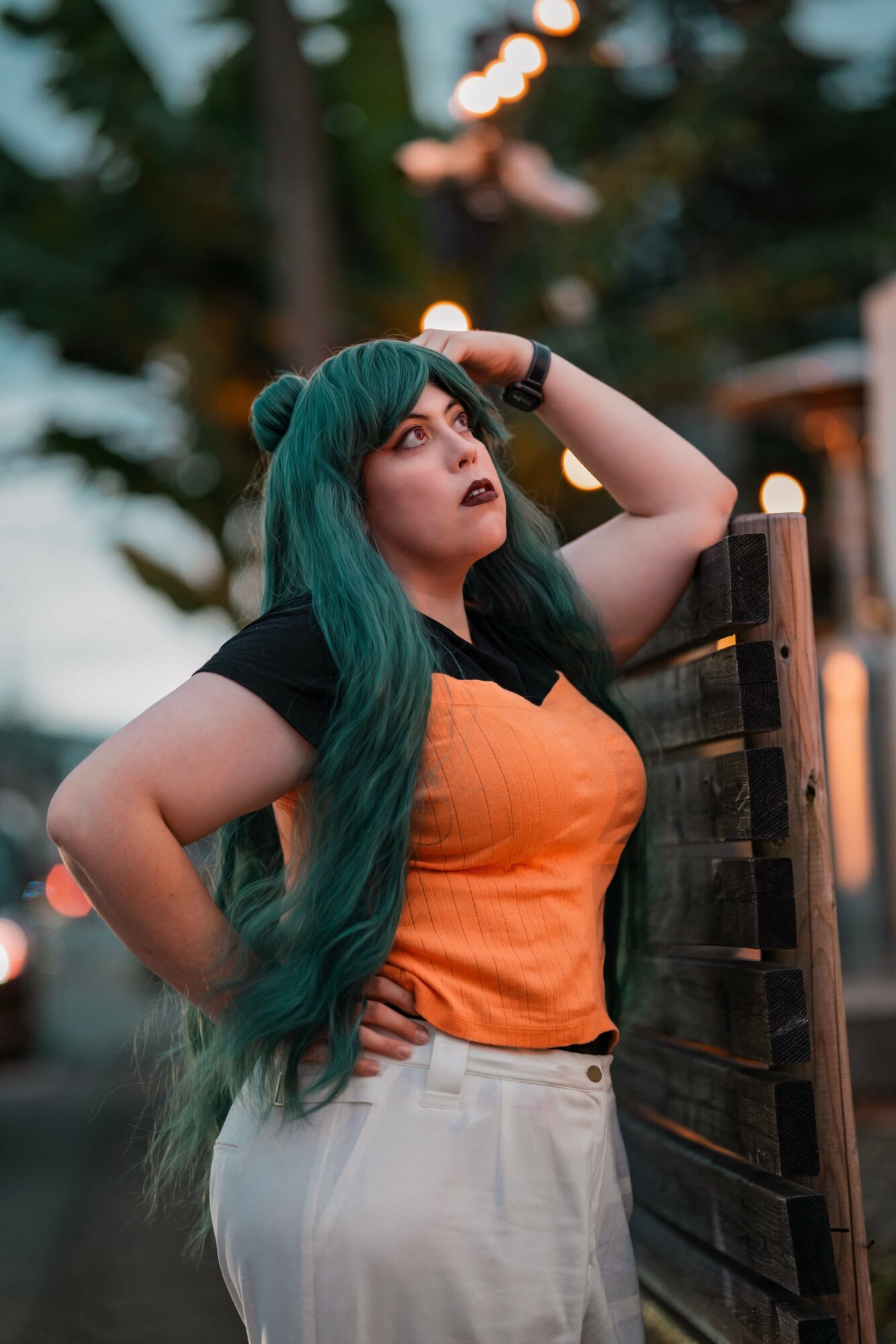Woman with long green hair wearing an orange top and white pants, standing outdoors near a wooden fence.