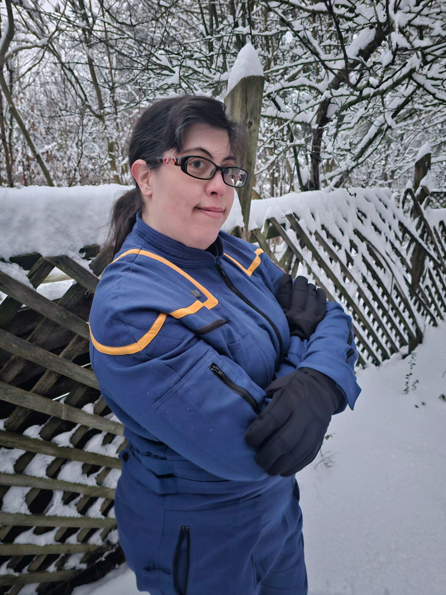 Person wearing glasses and a blue winter jacket standing outdoors in snow, with a wooden fence and snow-covered trees in background.
