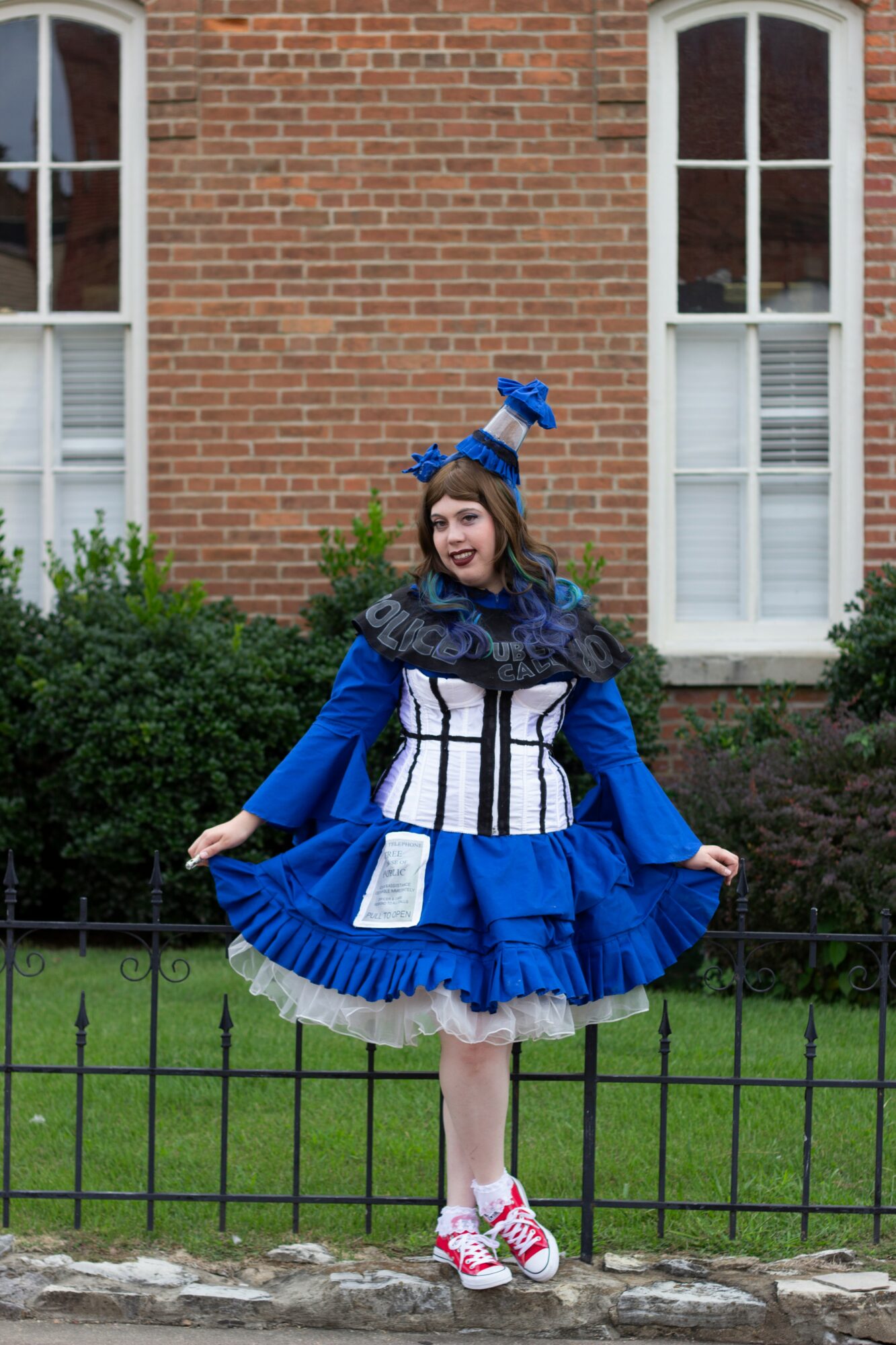 Girl in a blue and white costume with a matching headpiece, standing outdoors in front of a brick building and greenery.