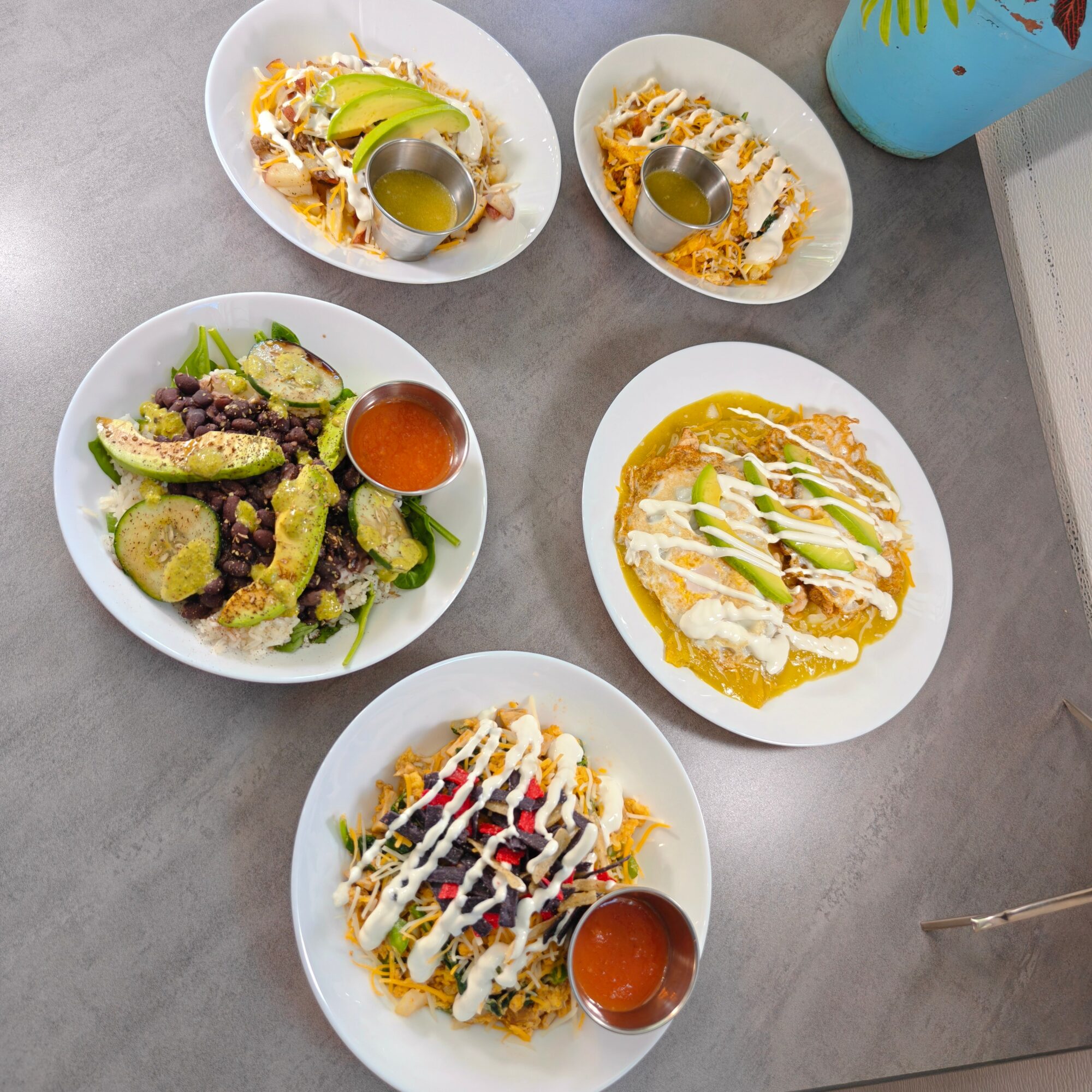 Five plates of food on a gray table, including tacos, nachos, and a salad, with a blue drink nearby.
