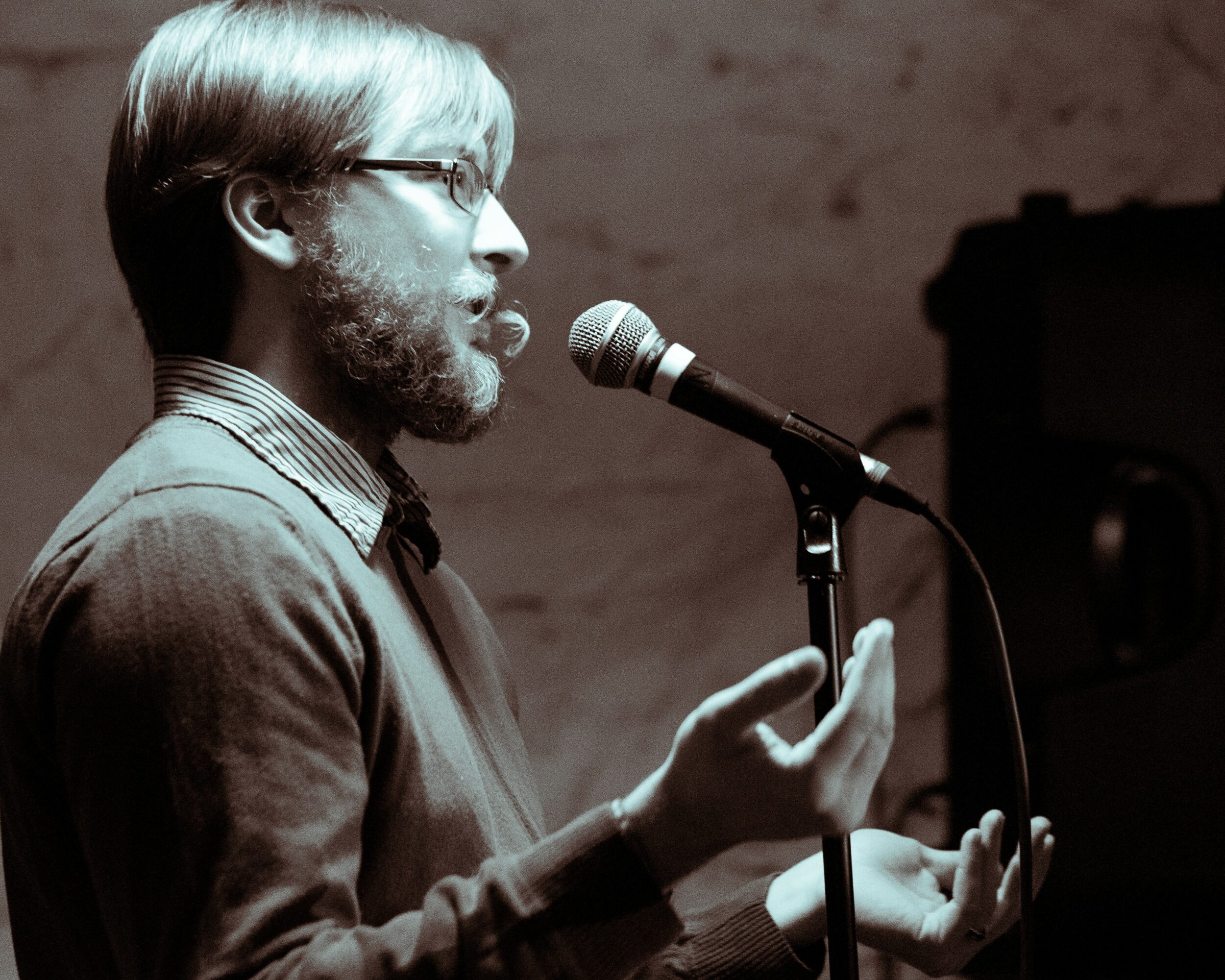 Person with glasses and beard speaking into a microphone, gesturing with hands, in a dimly lit room.