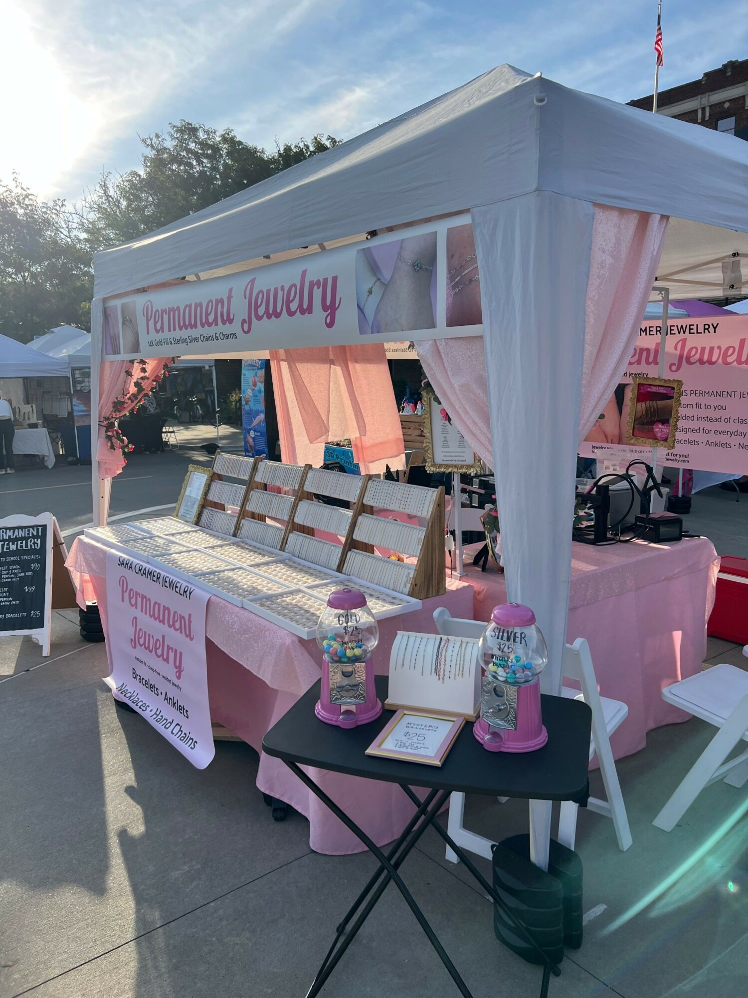Pink and white booth with products and signs, table with pink tablecloth, and chairs outside on pavement.