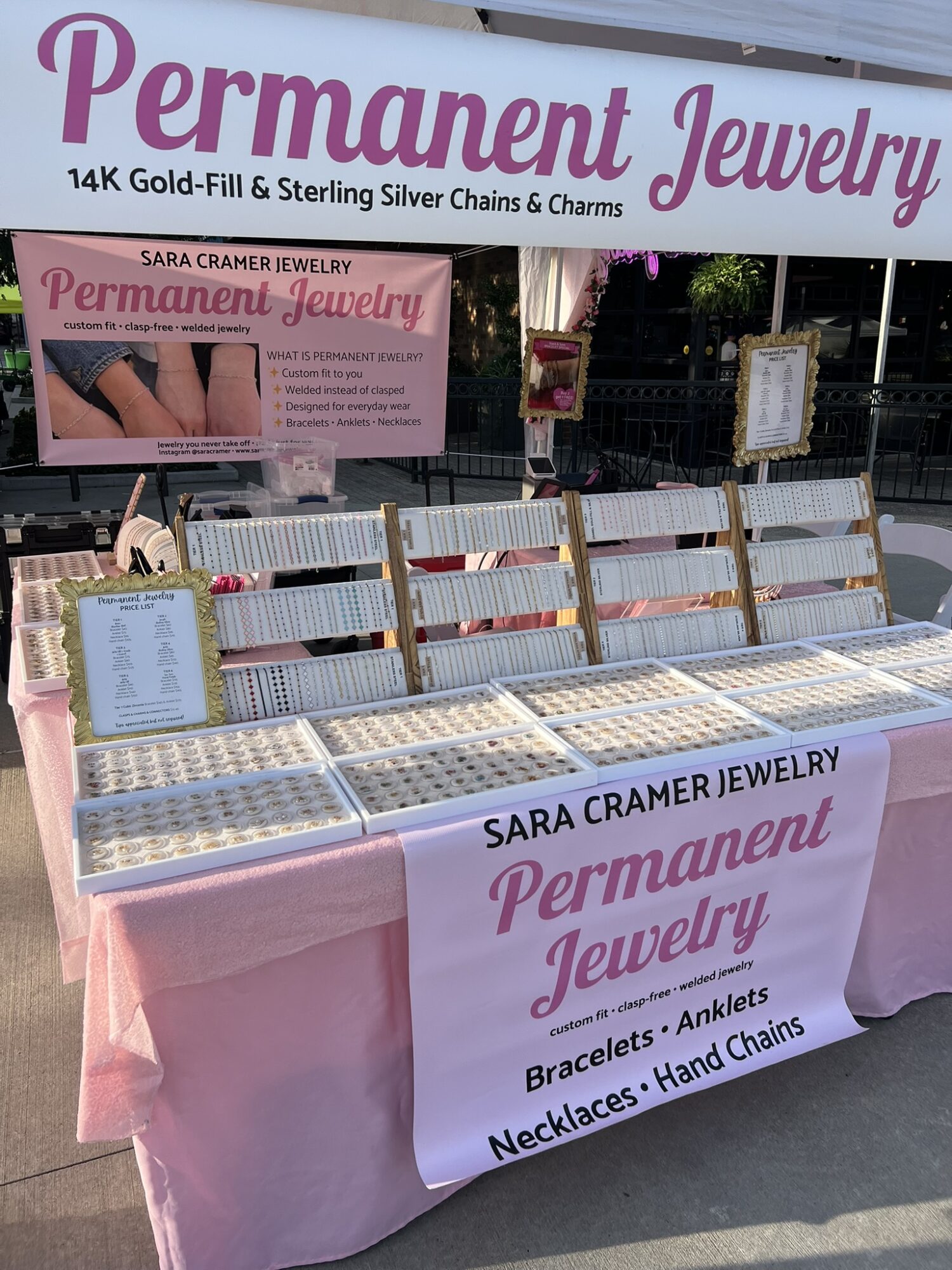 Jewelry display table with signs advertising permanent jewelry, including bracelets, anklets, necklaces, and hand chains, at an outdoor market.