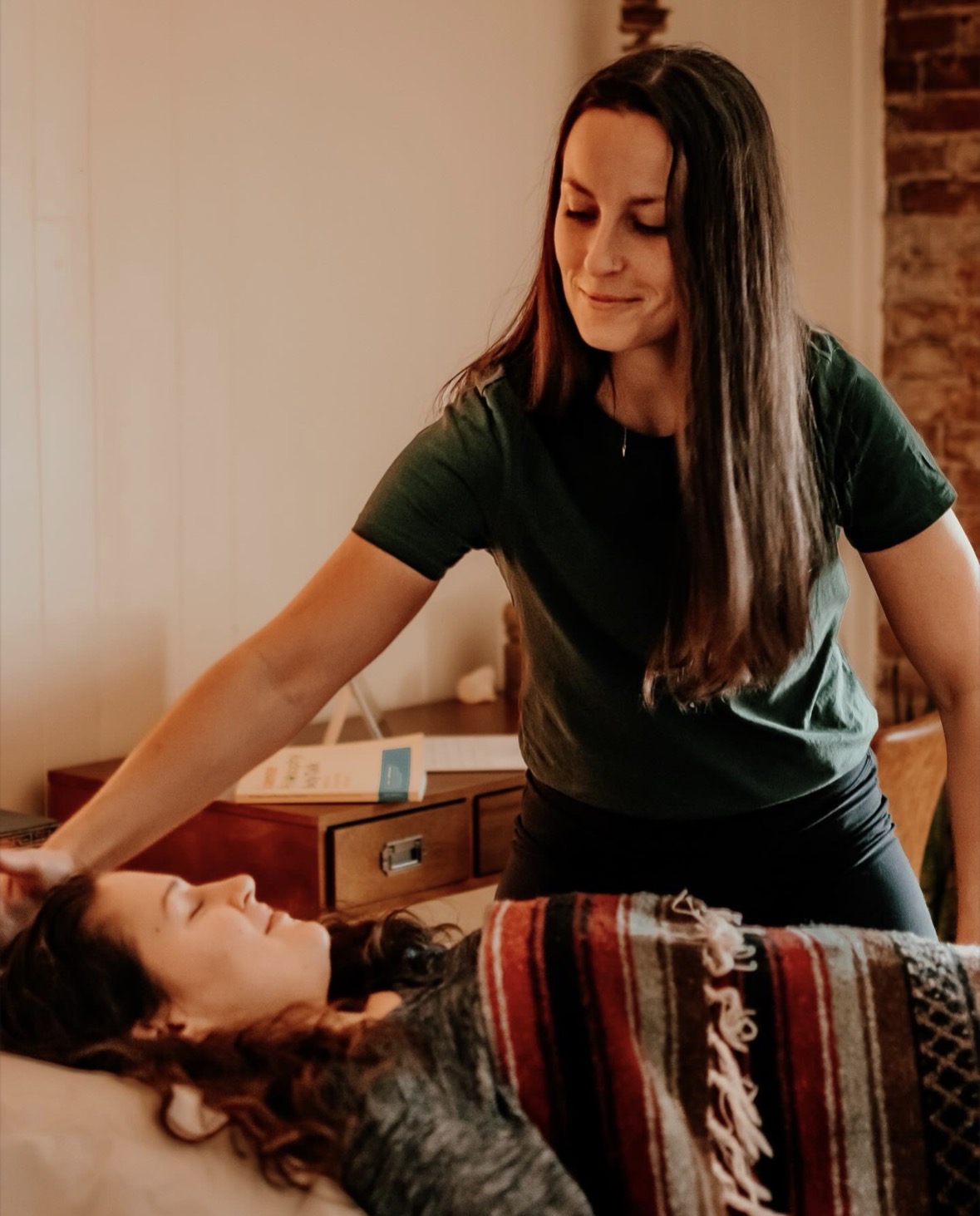 Woman leaning over a person lying in bed, in a room with a brick wall and wooden furniture.