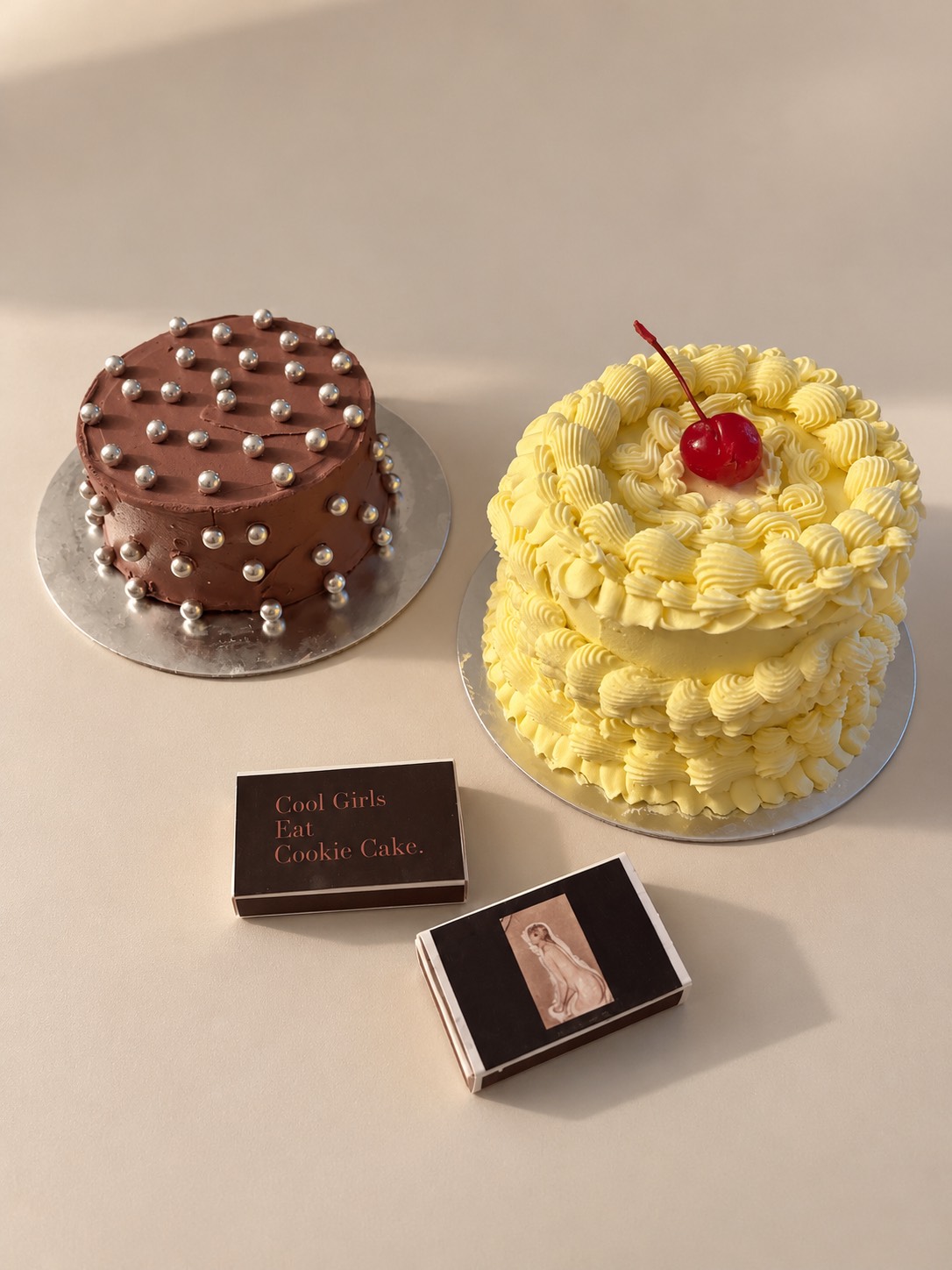 Two decorated cakes, one with chocolate and silver pearls, the other with yellow frosting and a cherry, on a white surface.