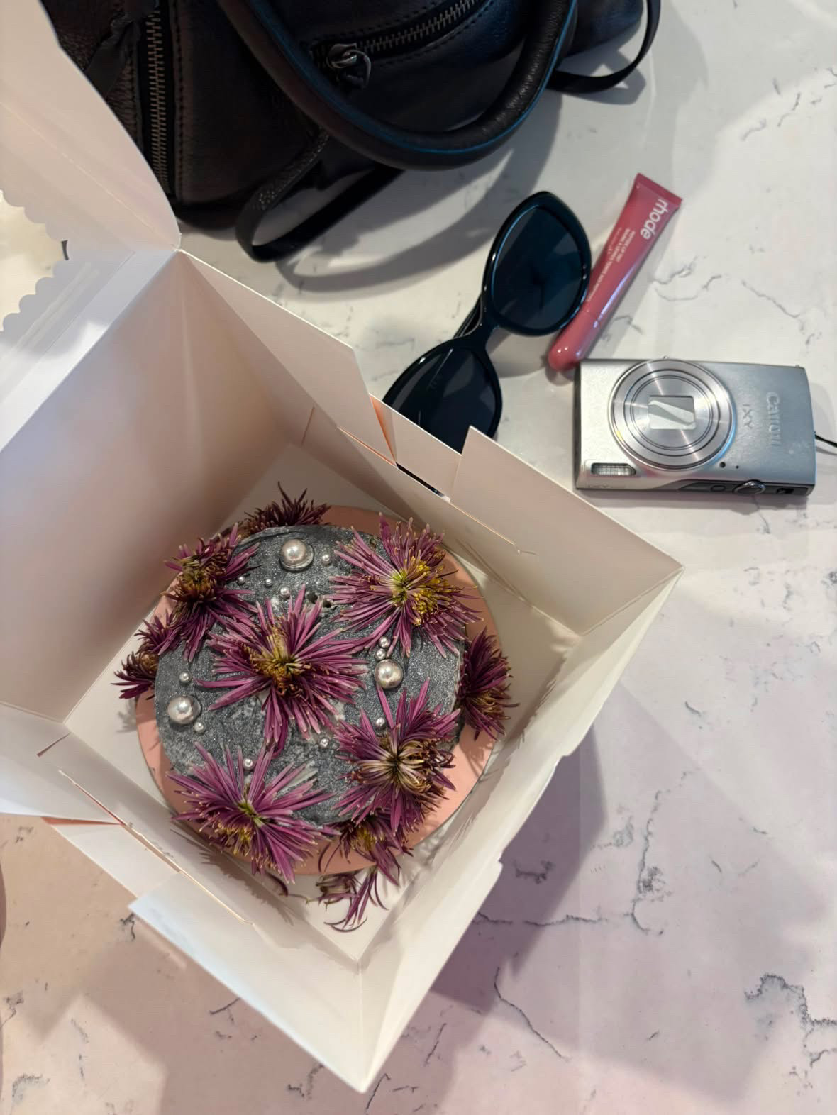 Pink fabric-covered item with purple and yellow flowers and pearl accents inside a white box on a marble surface.
