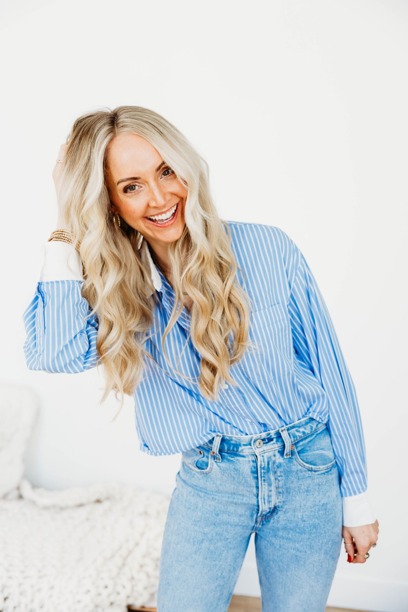 Woman with long wavy blonde hair smiling, wearing a blue striped shirt and jeans, standing against a white background.