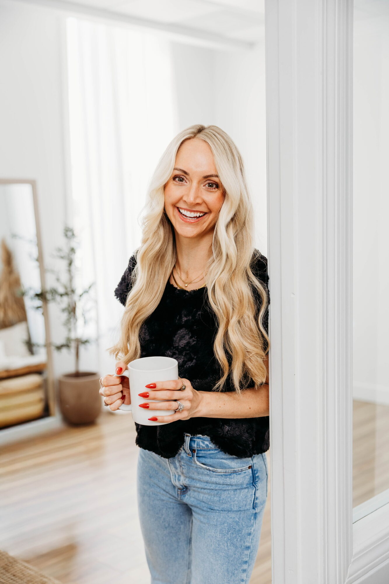 Woman with long blonde hair smiling, holding a white mug, standing indoors near a white wall and window.
