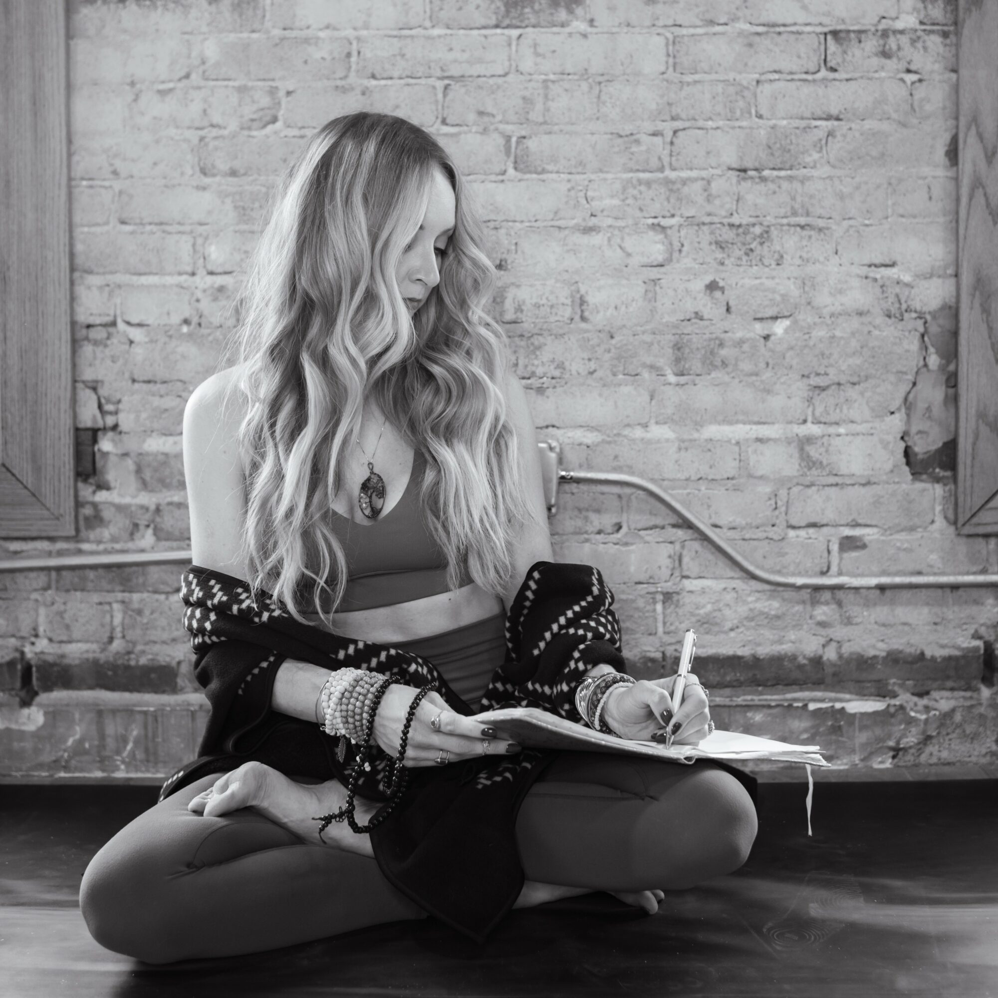 Woman with long wavy hair sitting cross-legged on the floor, writing in a notebook, against a brick wall background.
