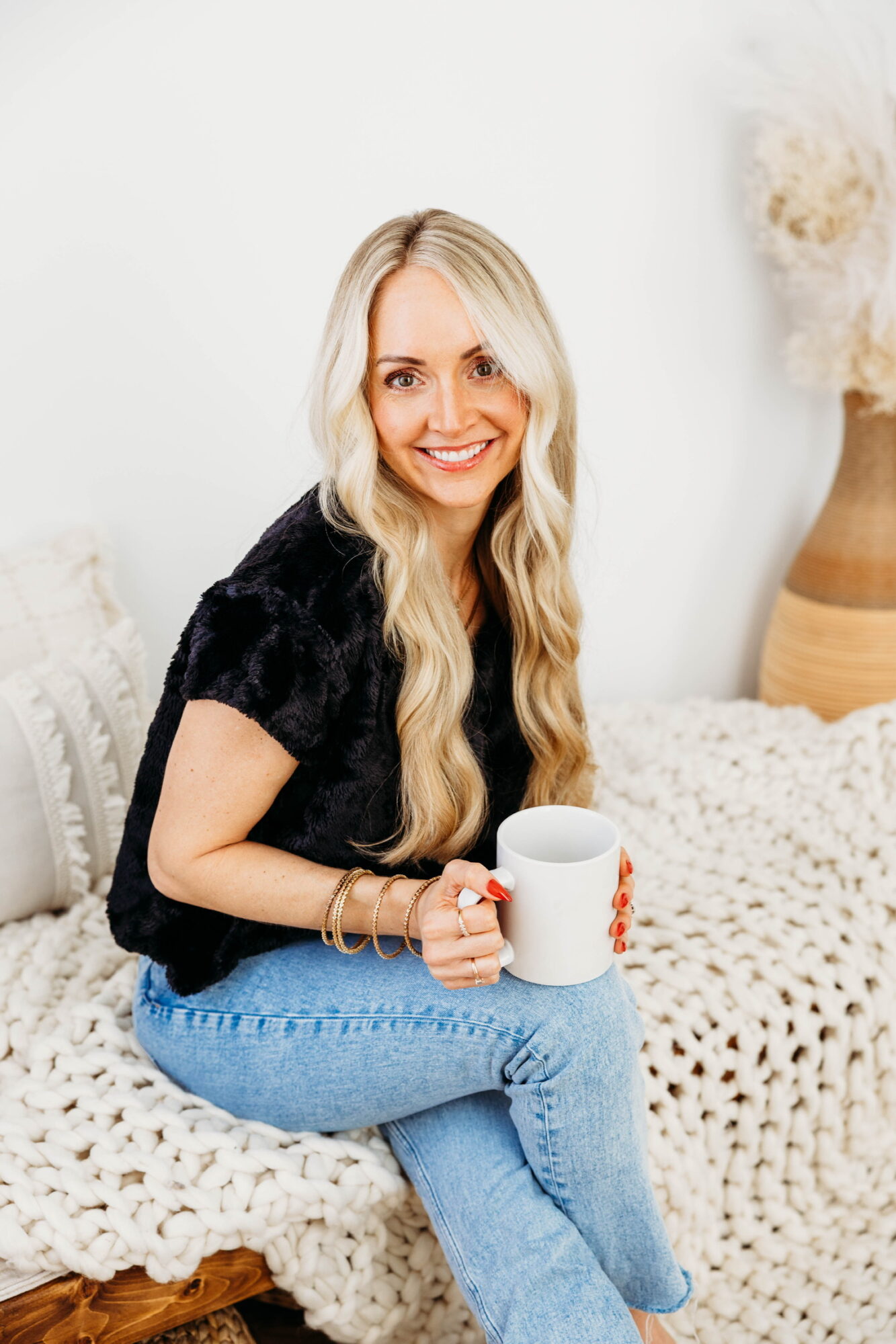 Woman with long blonde hair smiling, holding a white mug, sitting on a bed with a knitted blanket, in a bright room.