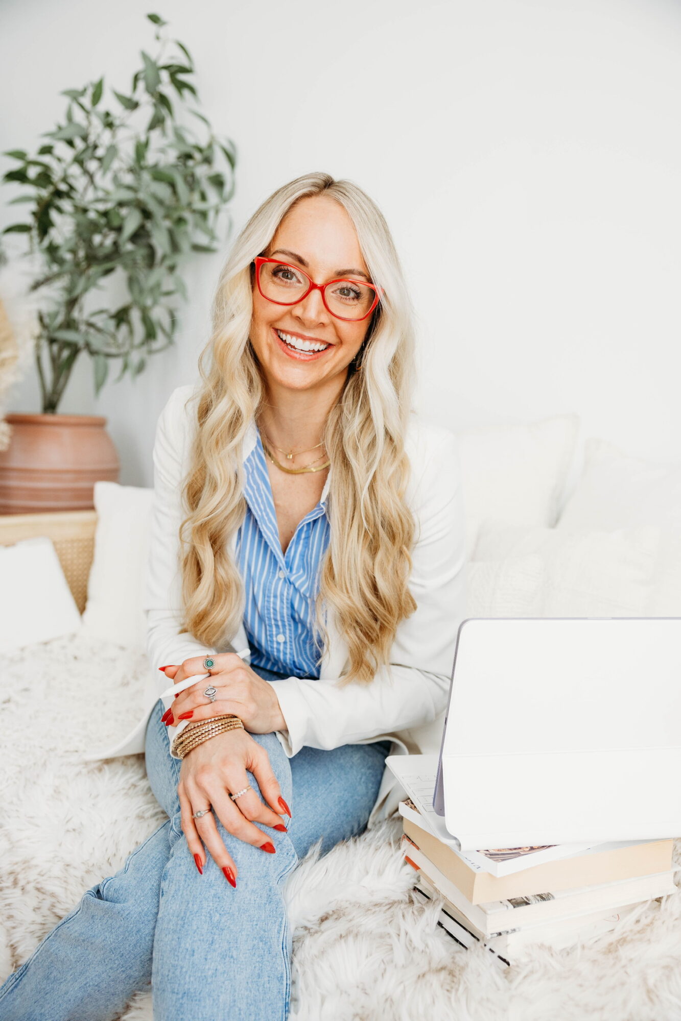 Smiling woman with long blonde hair and red glasses sitting on a couch with a laptop and books, in a bright room.