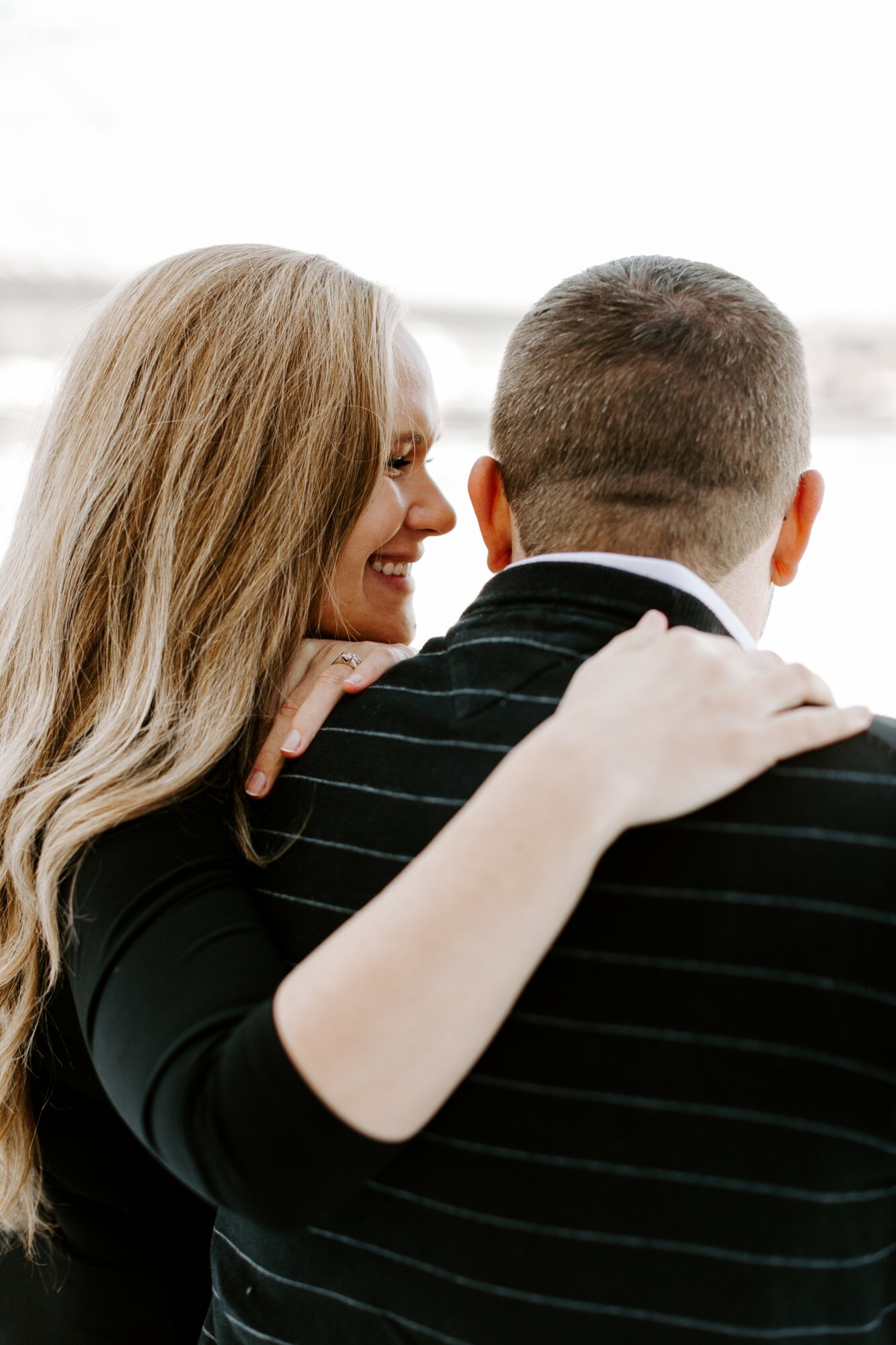 Woman with long blonde hair smiling, embracing a man with short hair, both facing away, indoors with bright background.