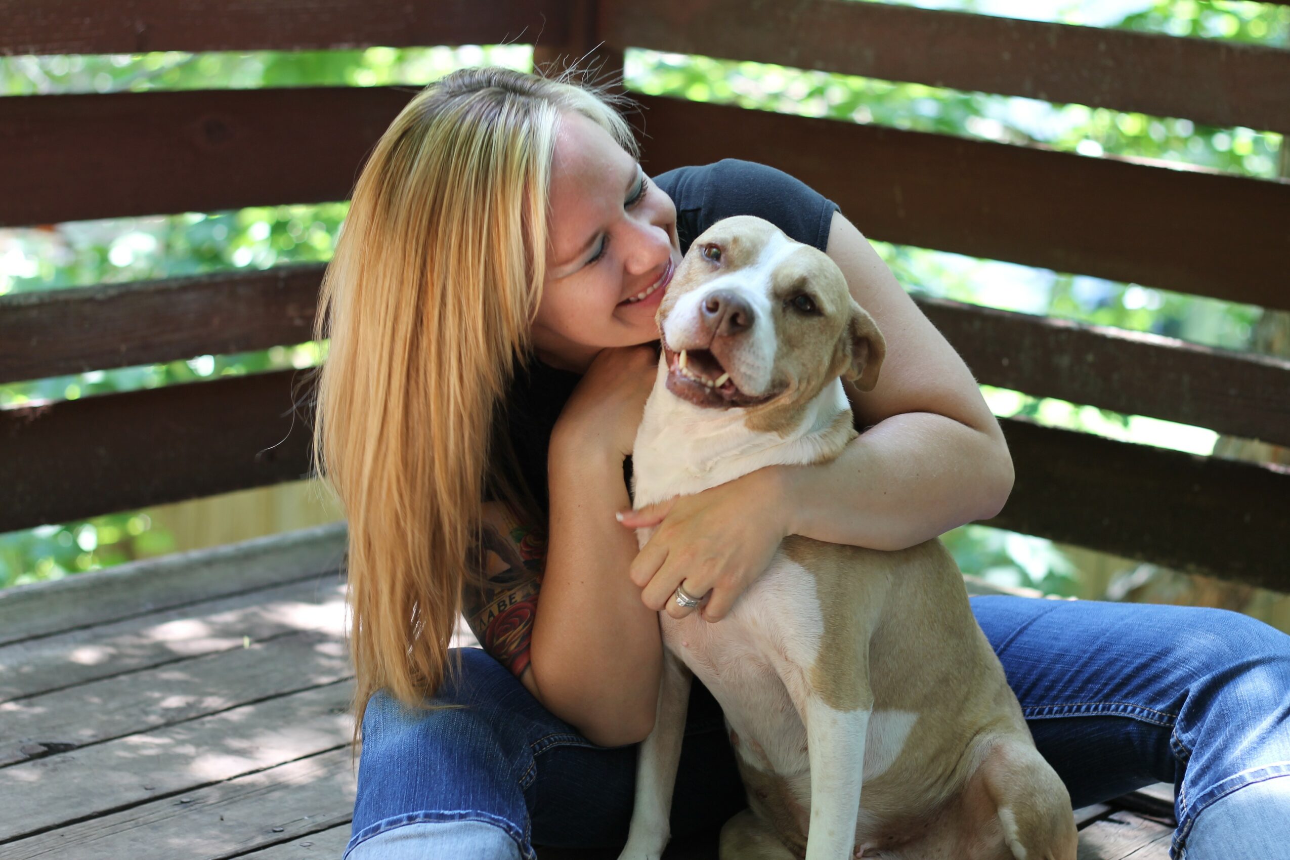 Girl hugging a smiling dog on a wooden deck with green foliage in the background.