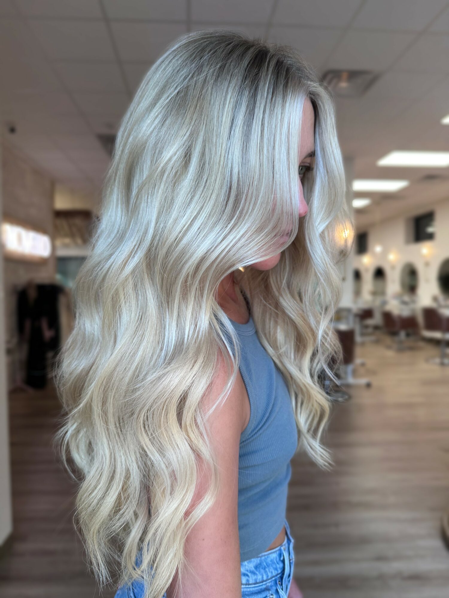 Side view of a woman with long, wavy platinum blonde hair in a salon, wearing a blue top.