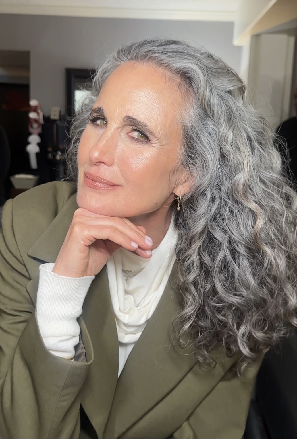 A woman with long, curly gray hair, wearing a green blazer and white scarf, resting her chin on her hand.