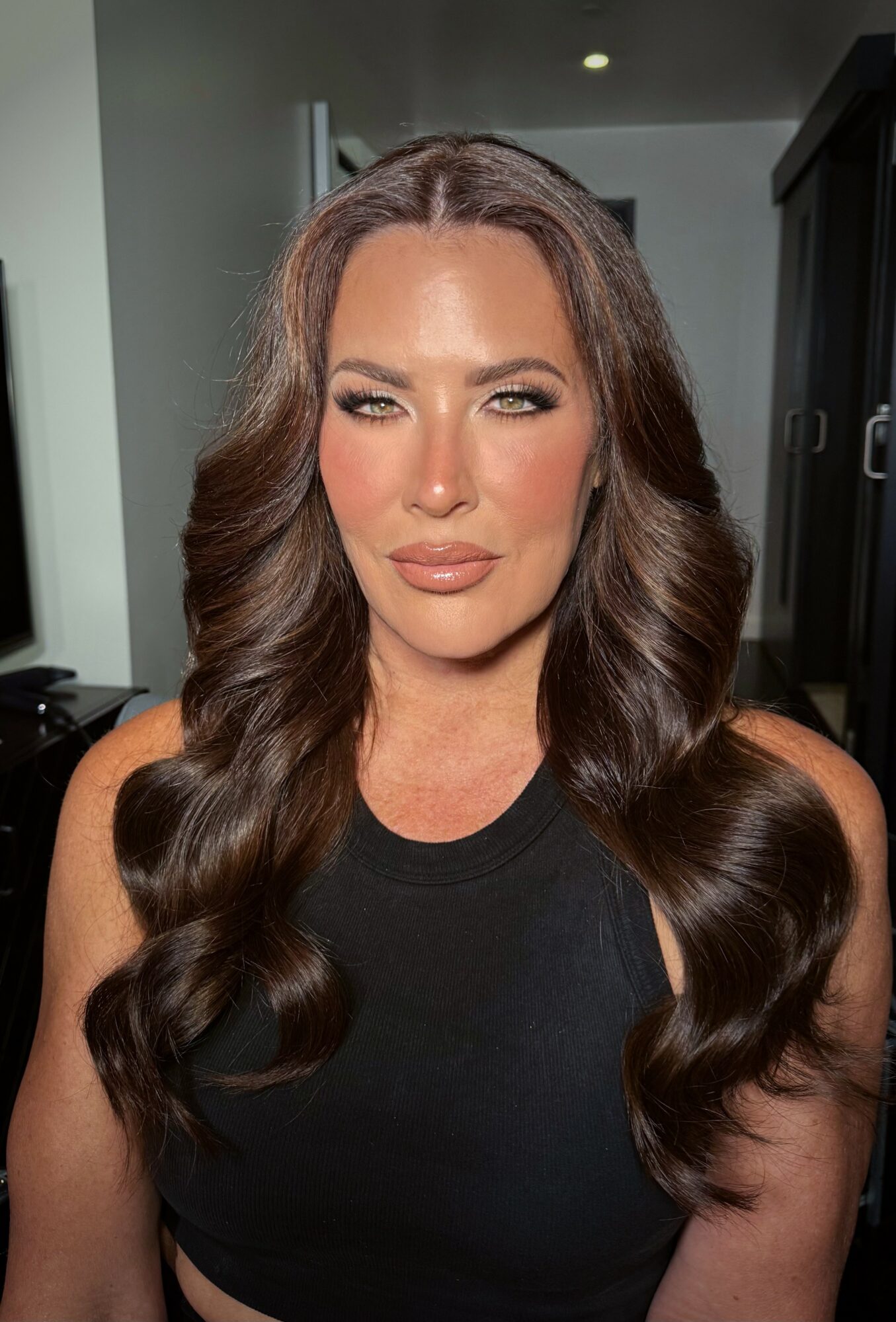 Woman with long, wavy brown hair and makeup, wearing a black sleeveless top, indoors with dark furniture in background.