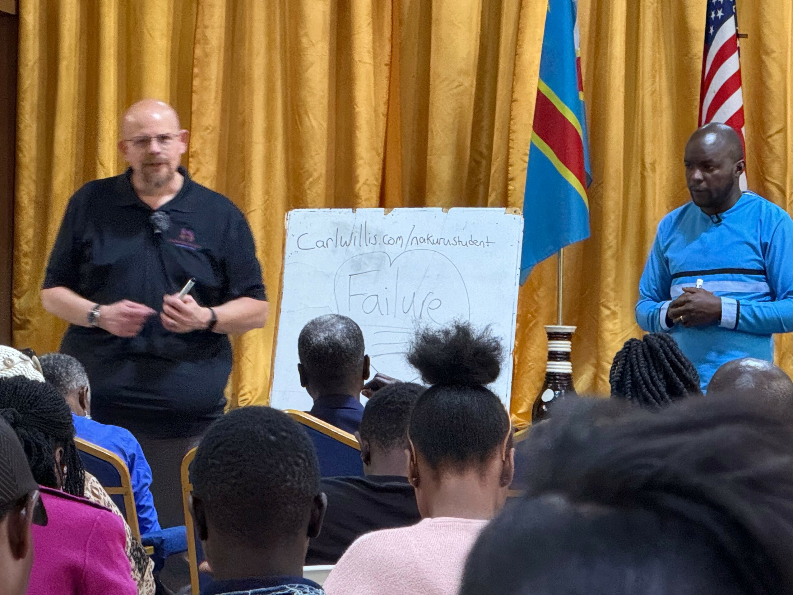 Two men stand in front of a gold curtain and flags, speaking to an audience, with a whiteboard behind them.