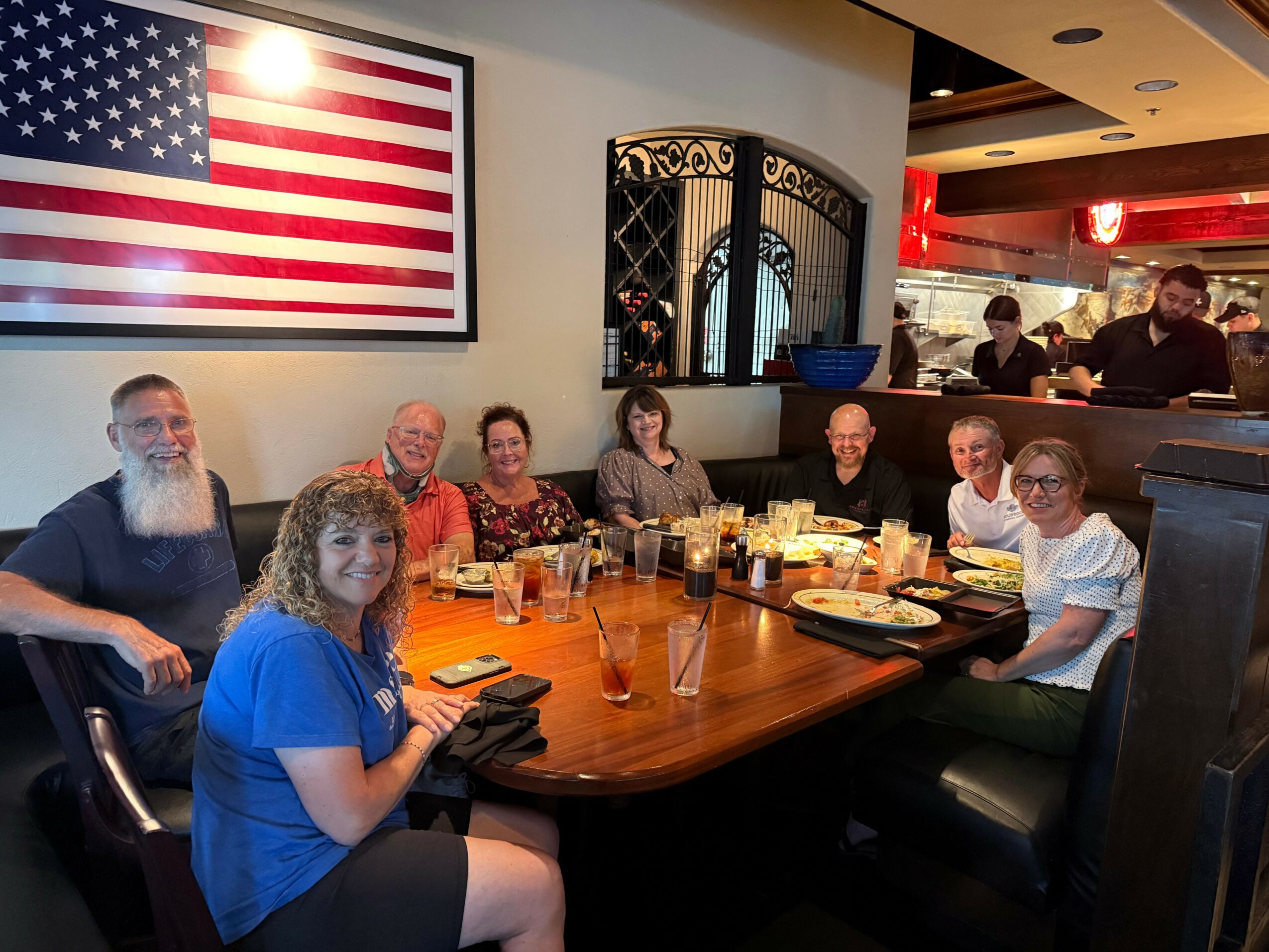 Group of nine people sitting around a restaurant table with food and drinks, American flag on wall behind them.