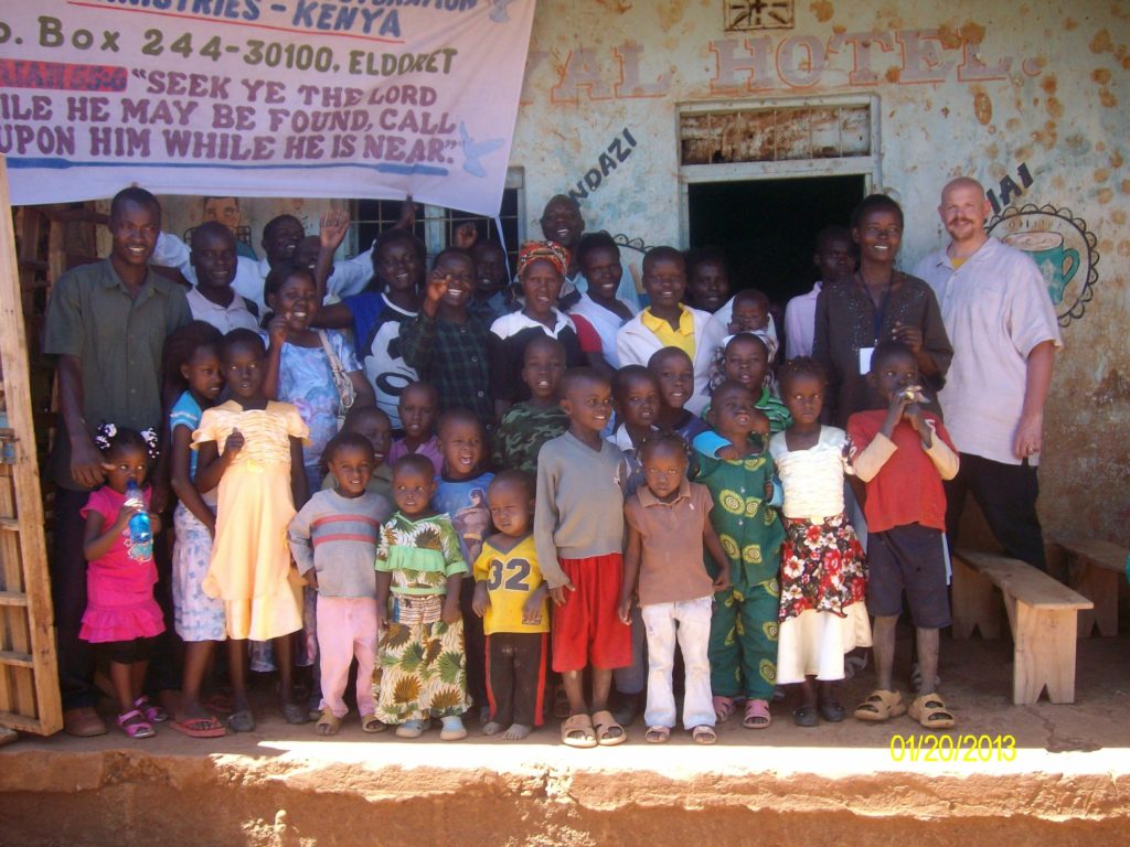 Group of children and adults standing outdoors in front of a wall with a banner, some children holding books or bags.