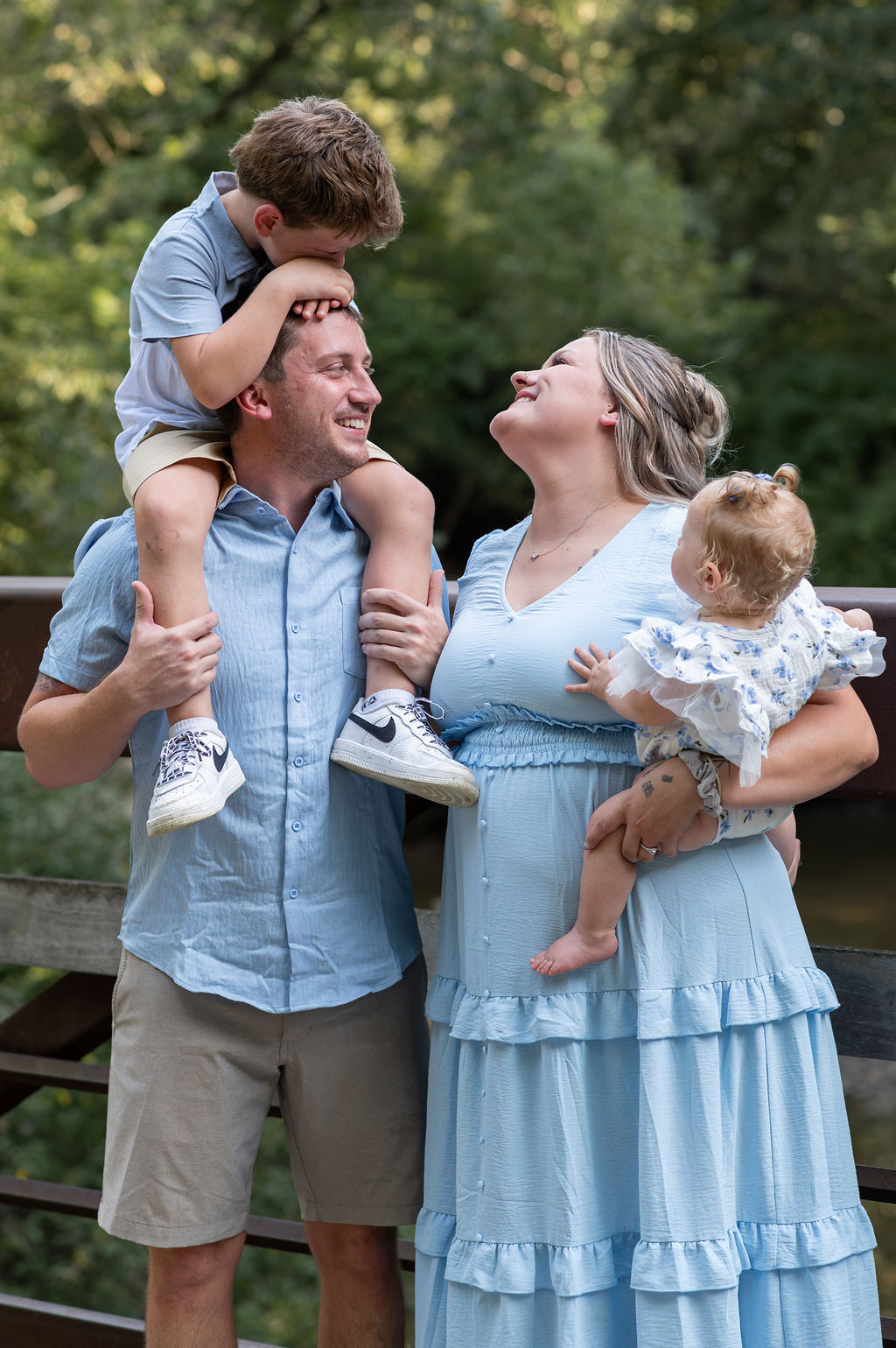 Family of four outdoors, father lifting son, mother holding daughter, smiling, trees in background