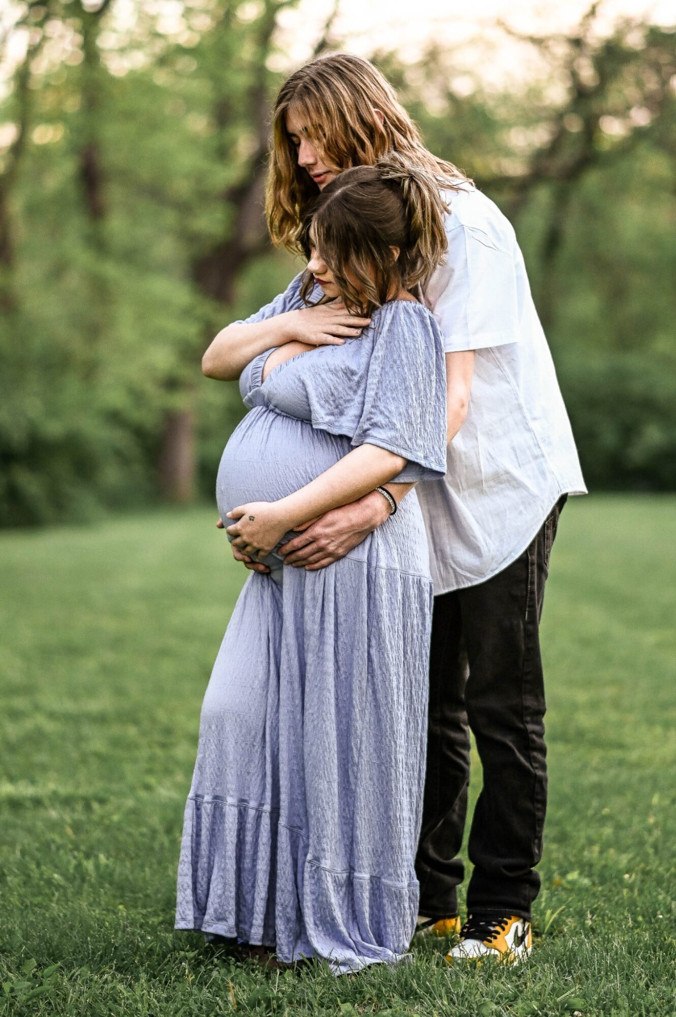 Two women, one pregnant, embrace outdoors in a grassy area with trees, one resting her head on the other's shoulder.