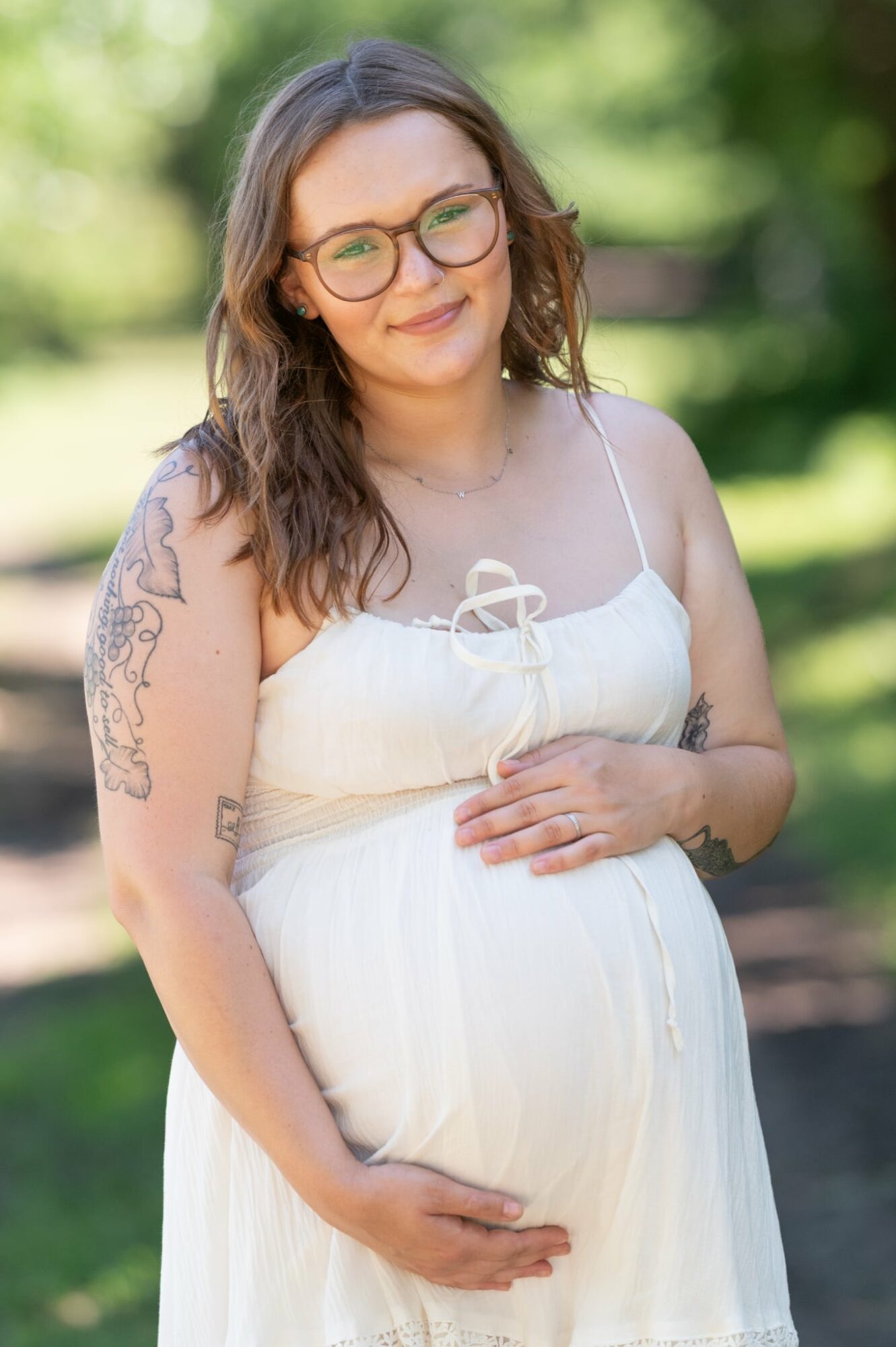Pregnant woman with glasses and tattoos, wearing a white dress, standing outdoors with greenery in background.
