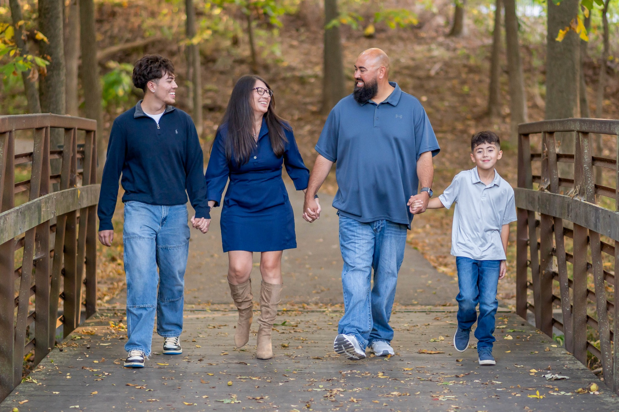 Family of four walking hand-in-hand on a wooden bridge in a forested area, smiling and enjoying nature.