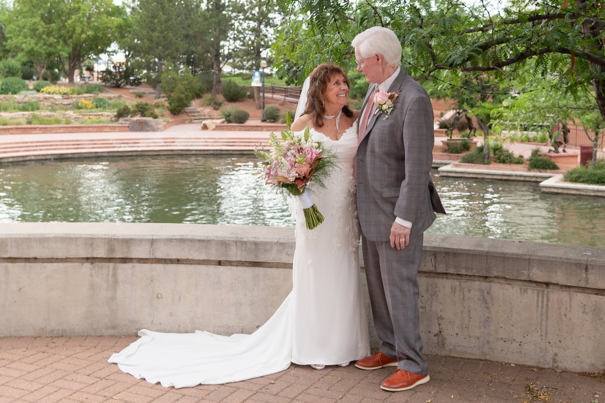 Bride and an older man stand by a pond in a park, holding hands and looking at each other, surrounded by trees.