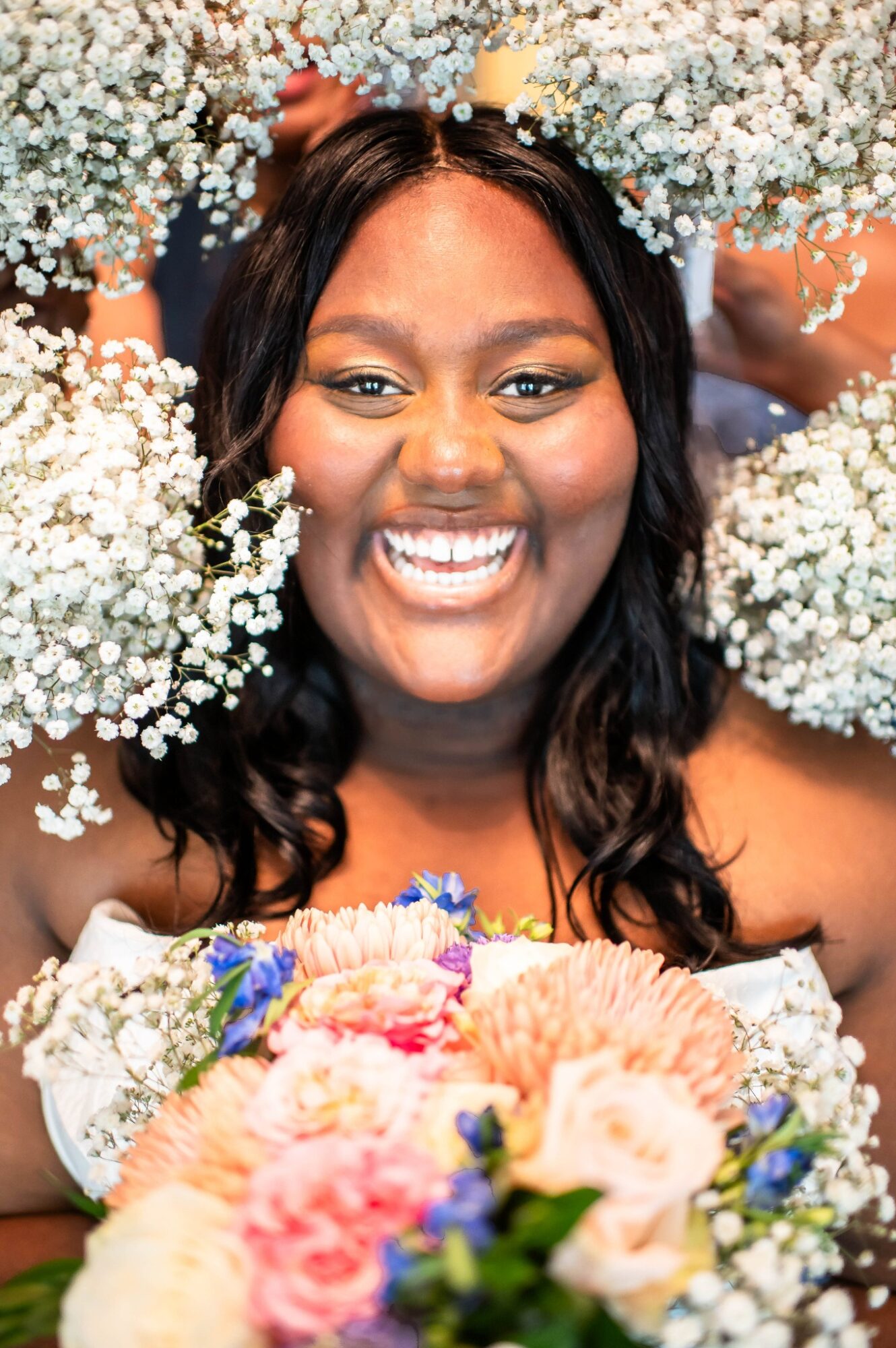 Smiling woman with dark hair surrounded by white flowers and a bouquet of pink, peach, and purple flowers.