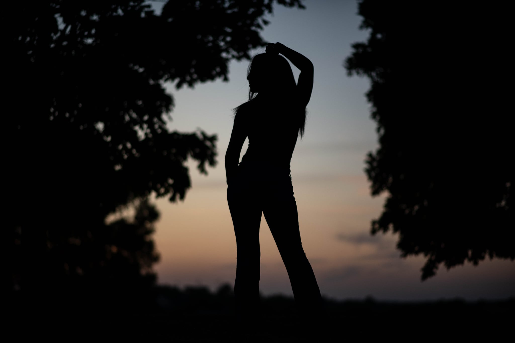 Silhouette of a person standing outdoors at sunset, with trees on either side and a colorful sky in the background.
