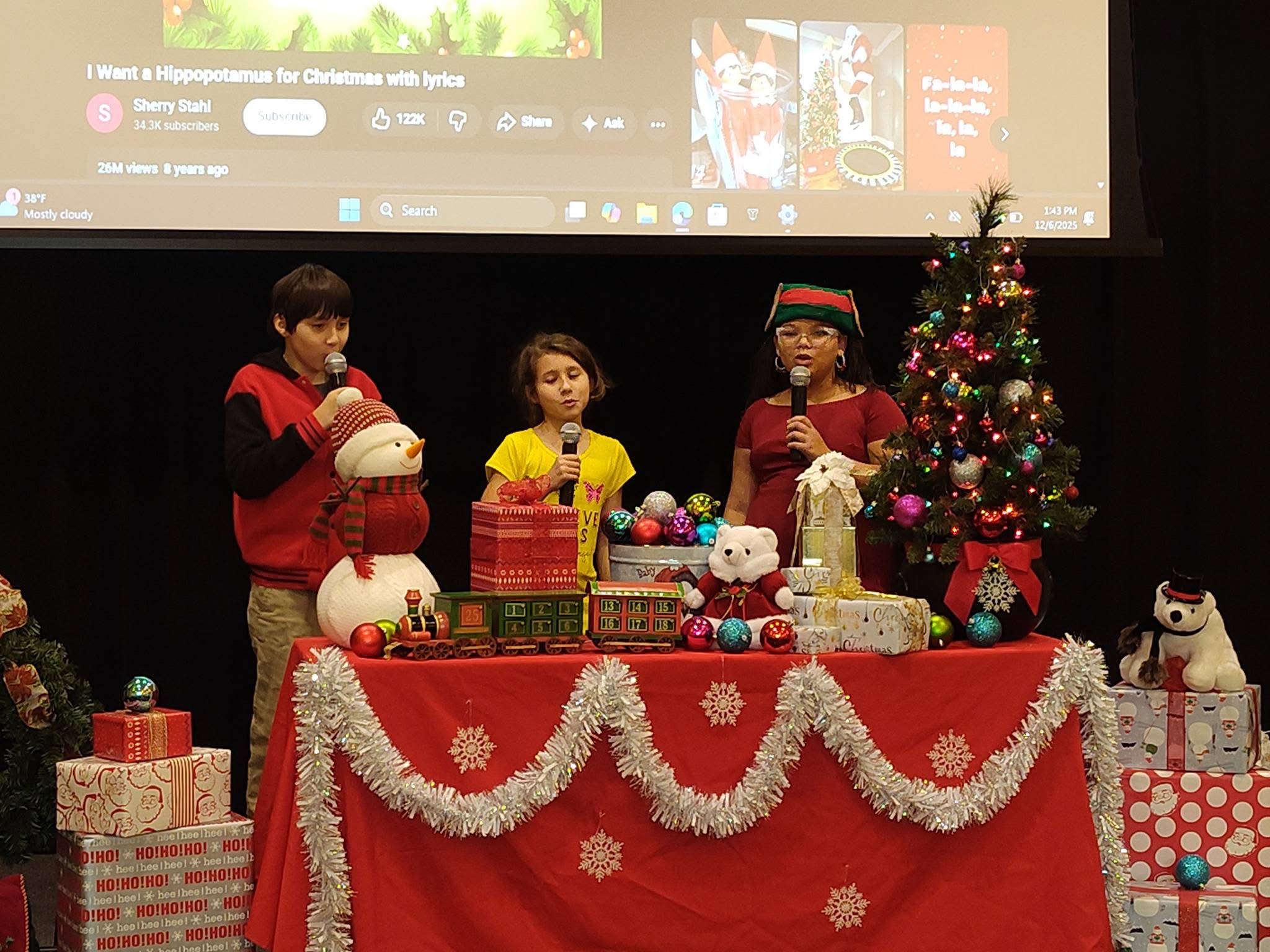 Three children stand behind a decorated table with Christmas presents and a tree, speaking into microphones during a holiday event.
