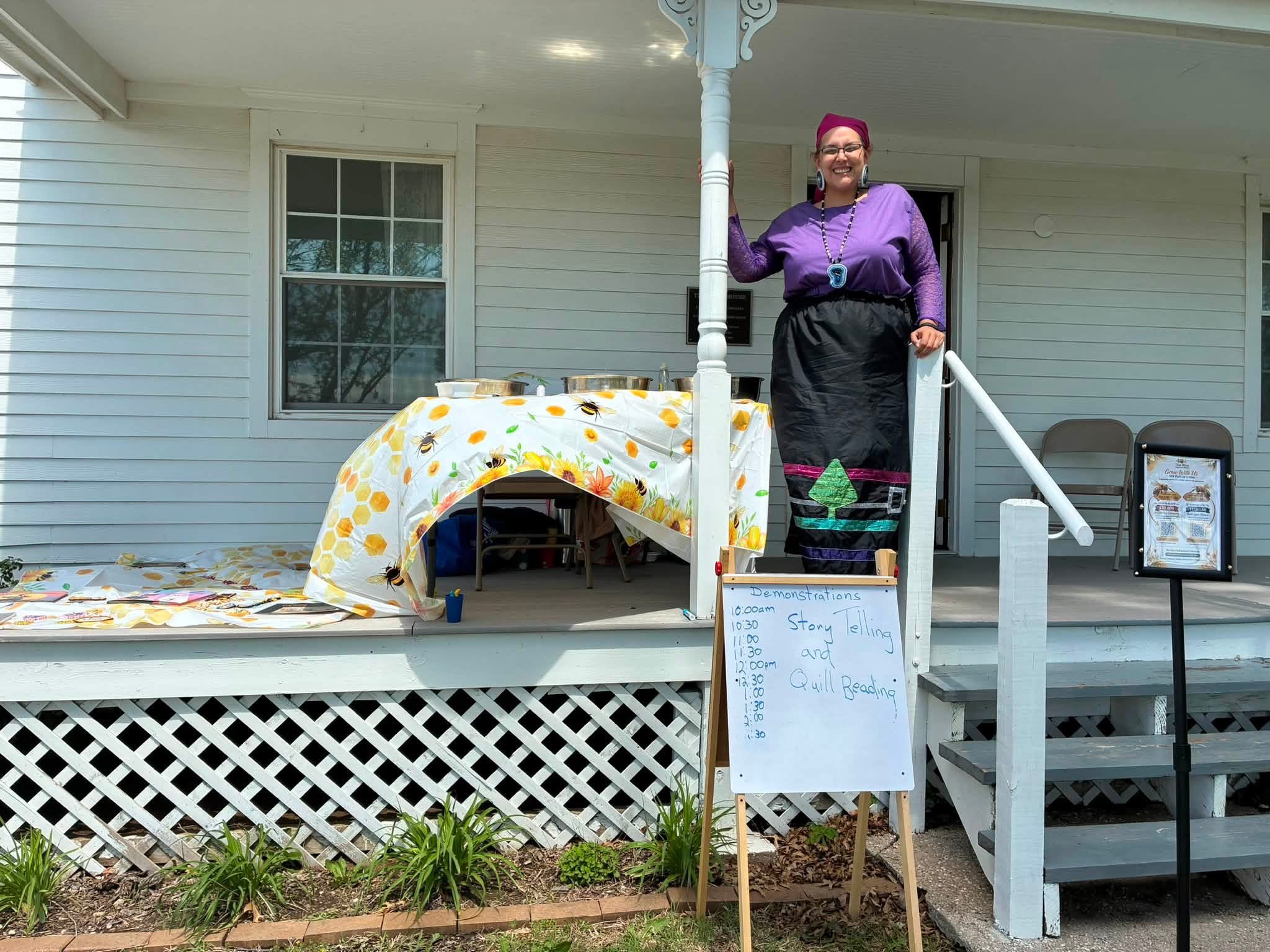 Woman standing on porch steps next to a table with a yellow polka dot tablecloth, in front of a white house with windows.
