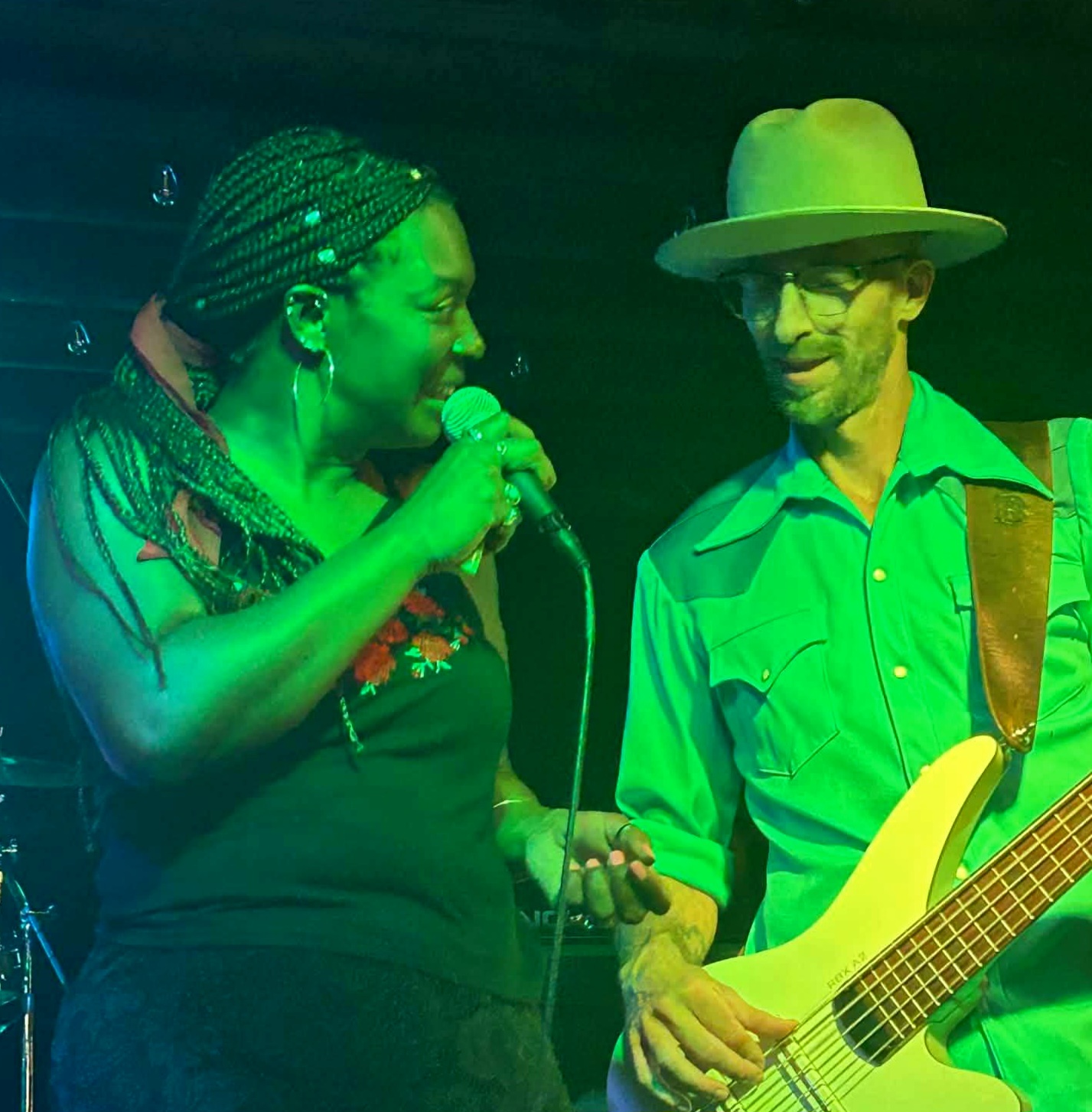 Two musicians perform on stage, one singing into a microphone and the other holding a bass guitar, both wearing colorful clothing.