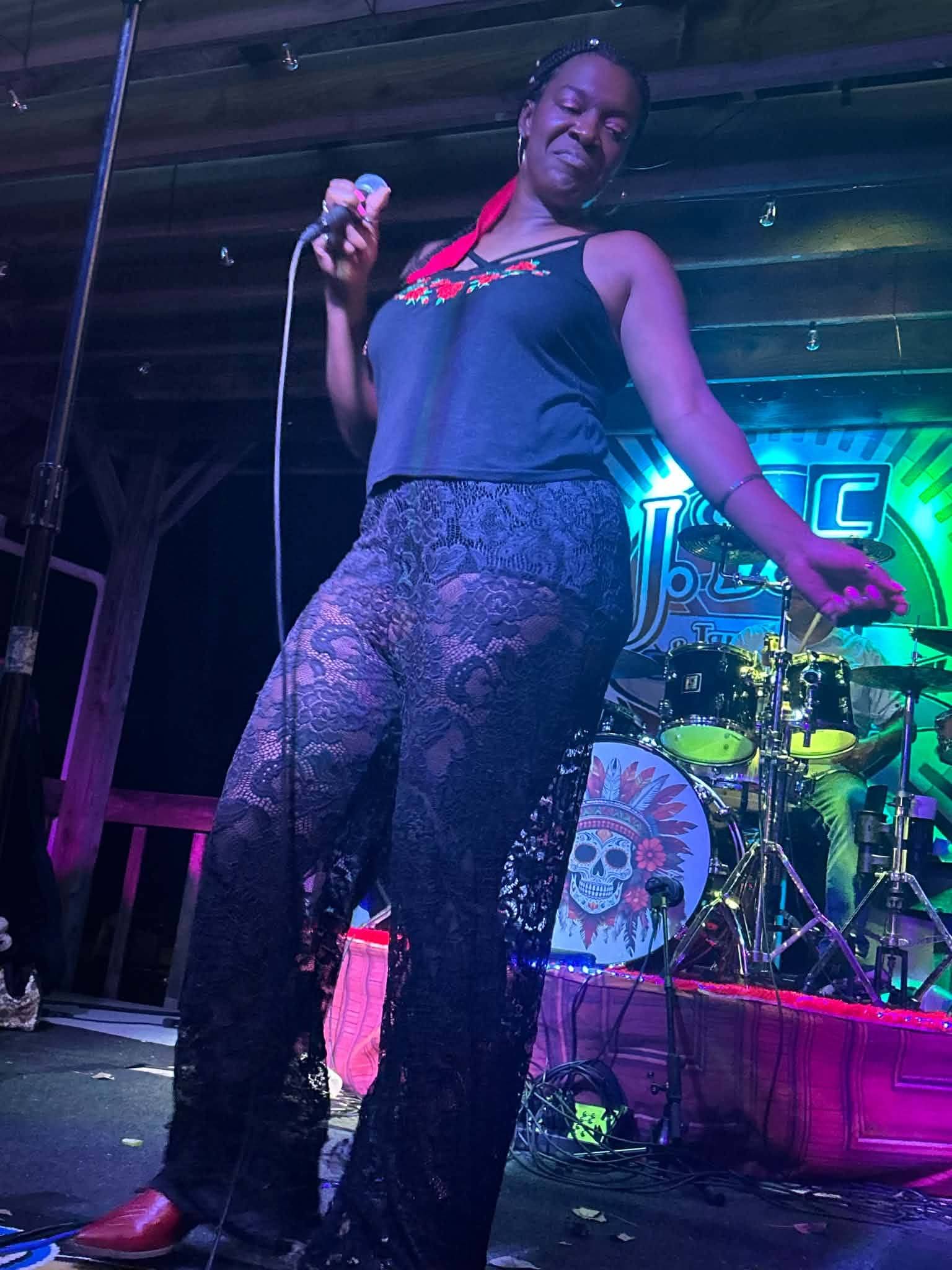 Performer singing on stage with a microphone, wearing a black top and patterned pants, smiling, with musical instruments in the background.