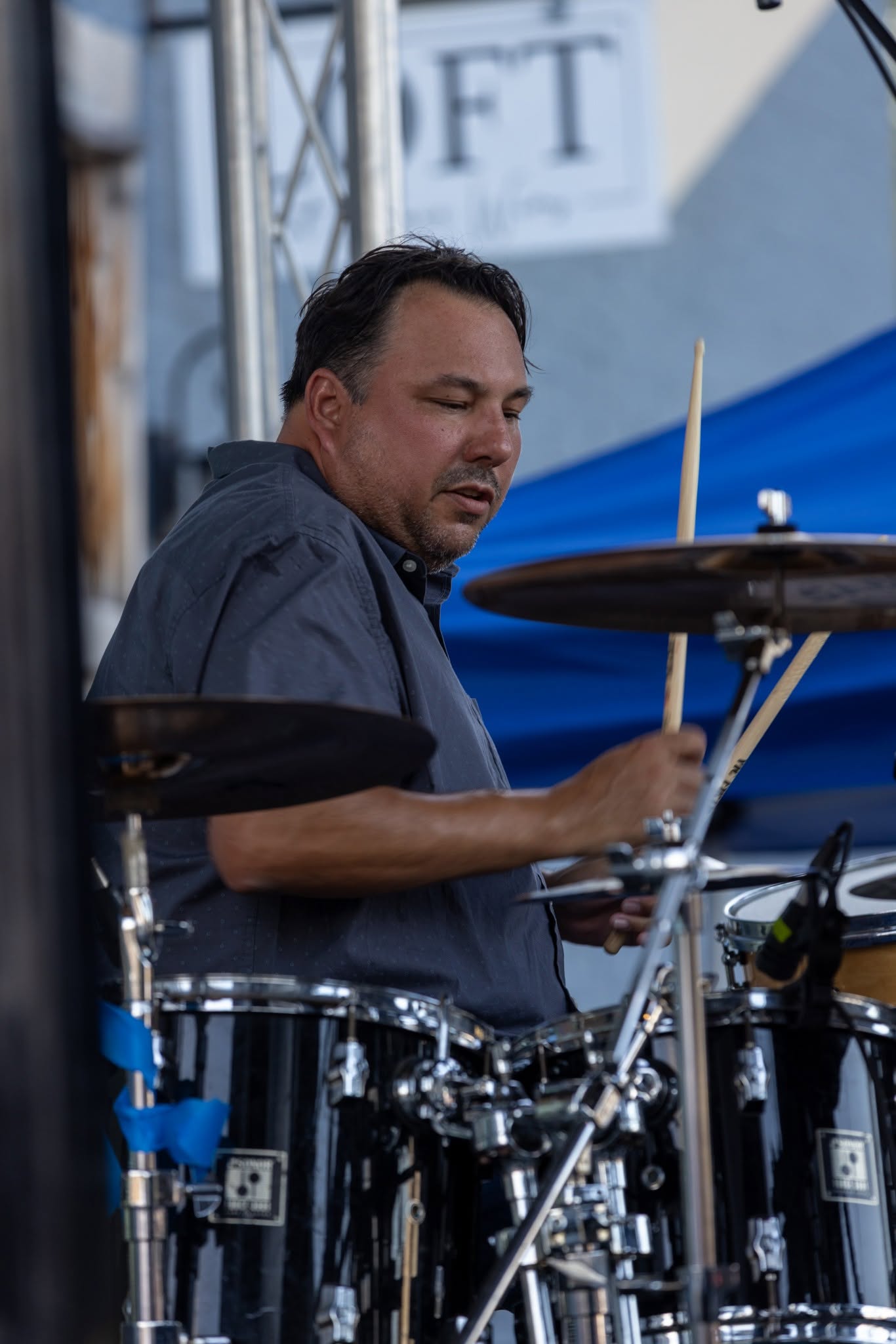 Man playing drums outdoors, focused on his instrument, with a blue tent and a sign in the background.