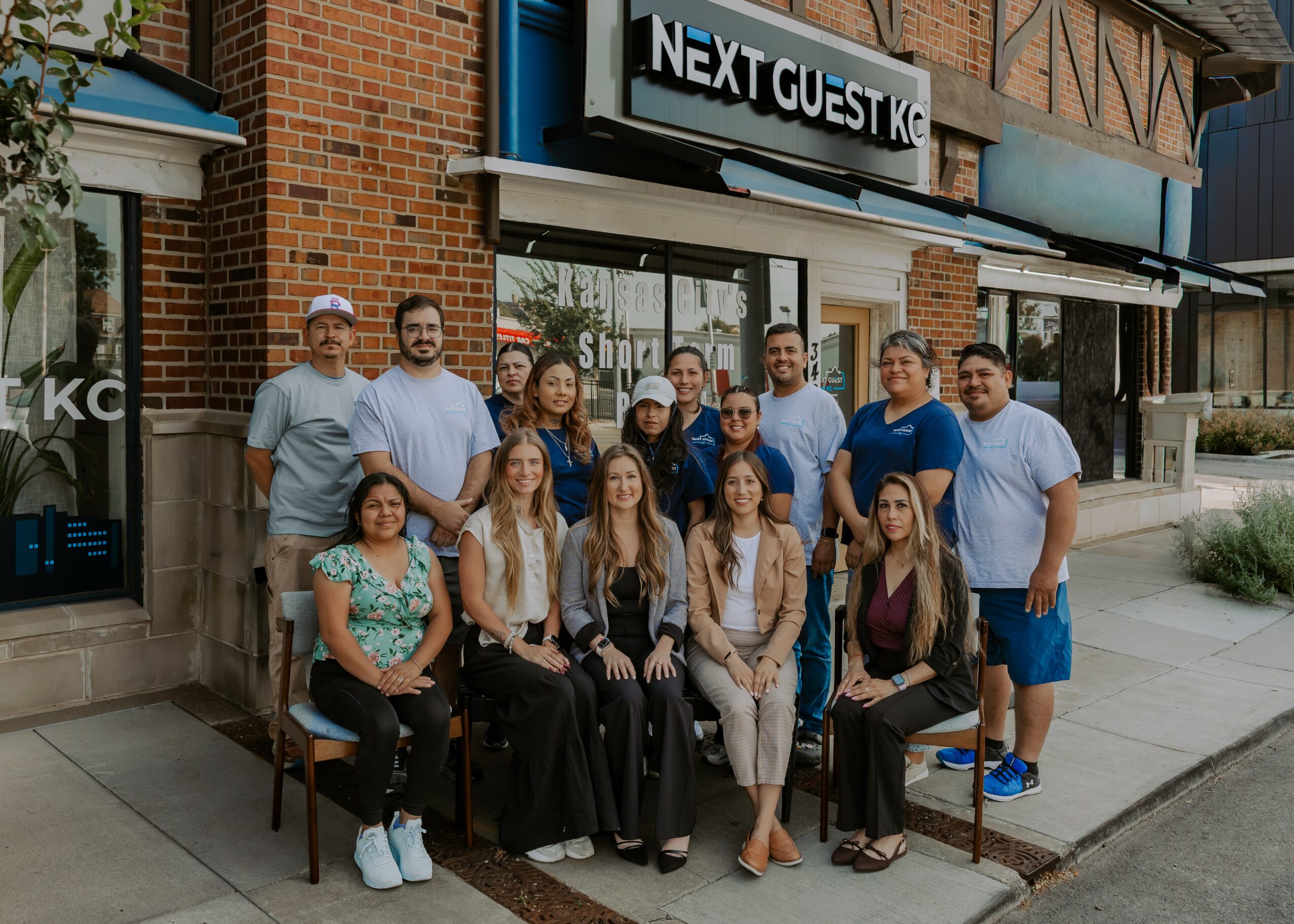 Group of people standing and sitting outside a building with a sign that reads 'Next Guest'.