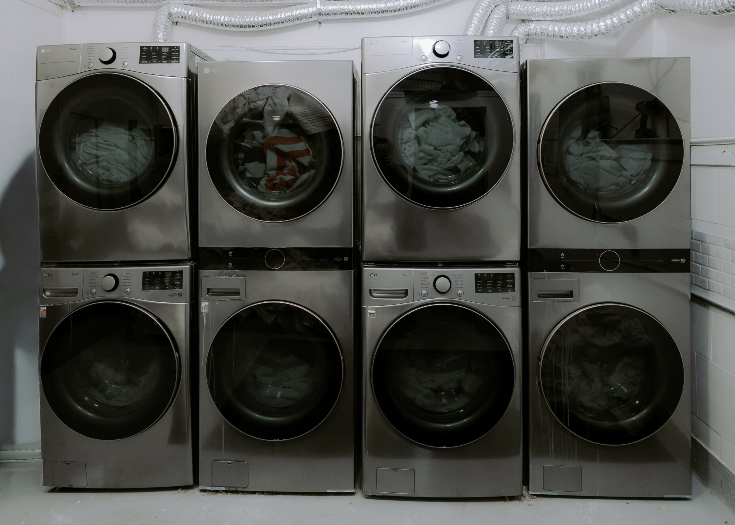 Eight front-loading washing machines stacked in two rows, with hoses connected at the top, in a laundry room.