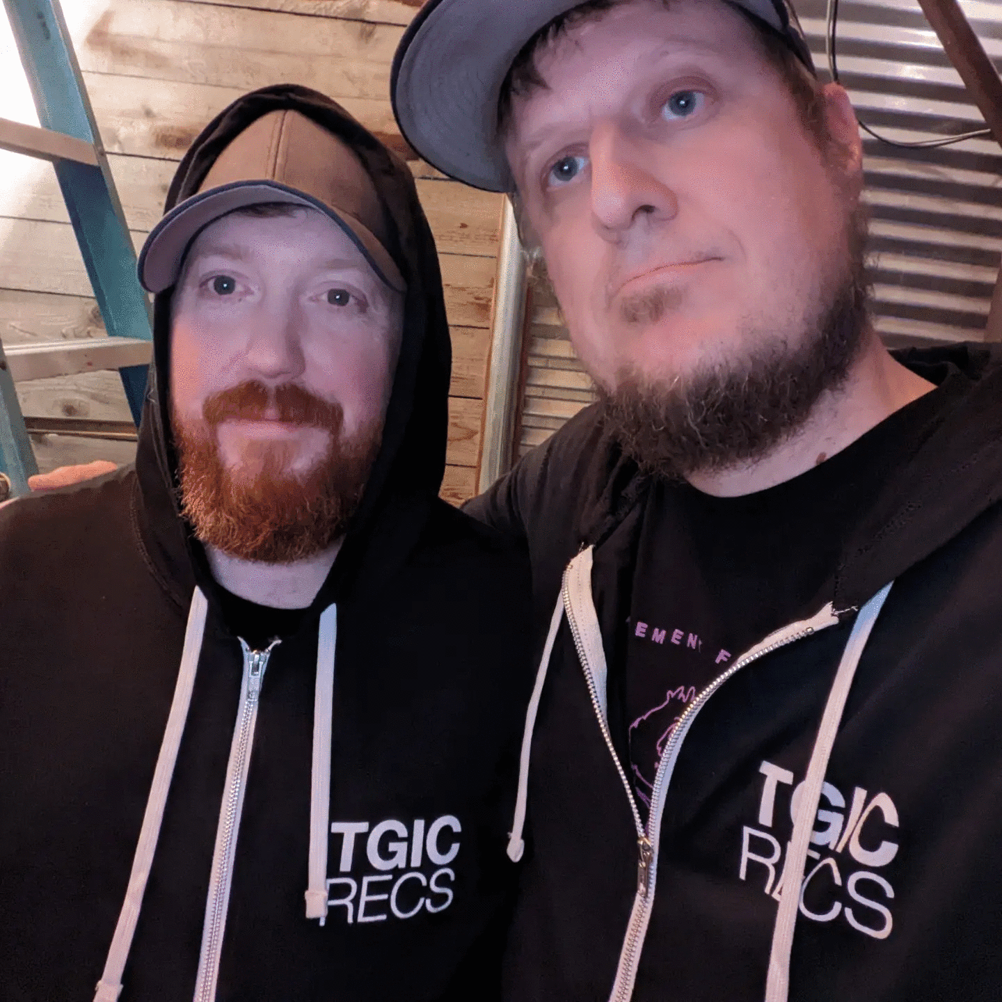 Two men with beards and caps, wearing black hoodies with white text, pose indoors with wooden and metal background.
