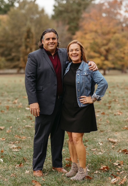 Two people standing outdoors on grass with trees in autumn background, smiling, one with arm around the other.