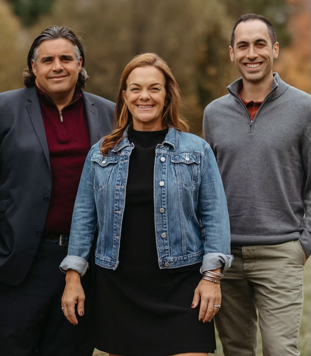Three people standing outdoors, smiling, with trees in the background.