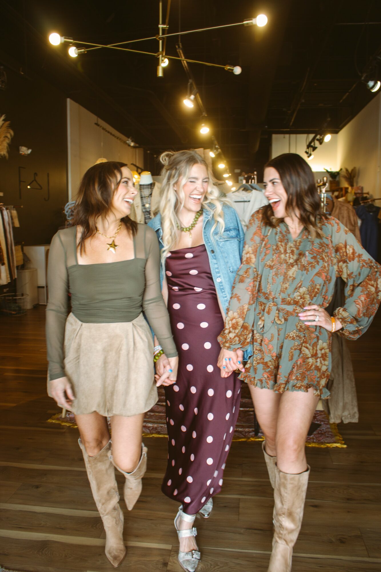 Three women smiling and holding hands in a store or event space with warm lighting and wooden floor.