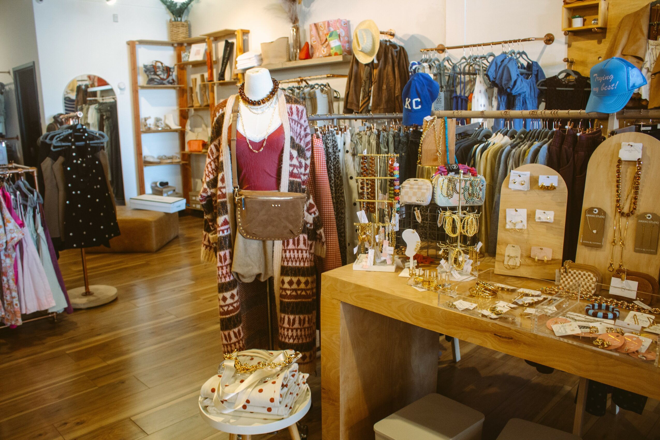 Clothing store with mannequins, shelves, and tables displaying accessories and apparel, wooden flooring, and organized displays.