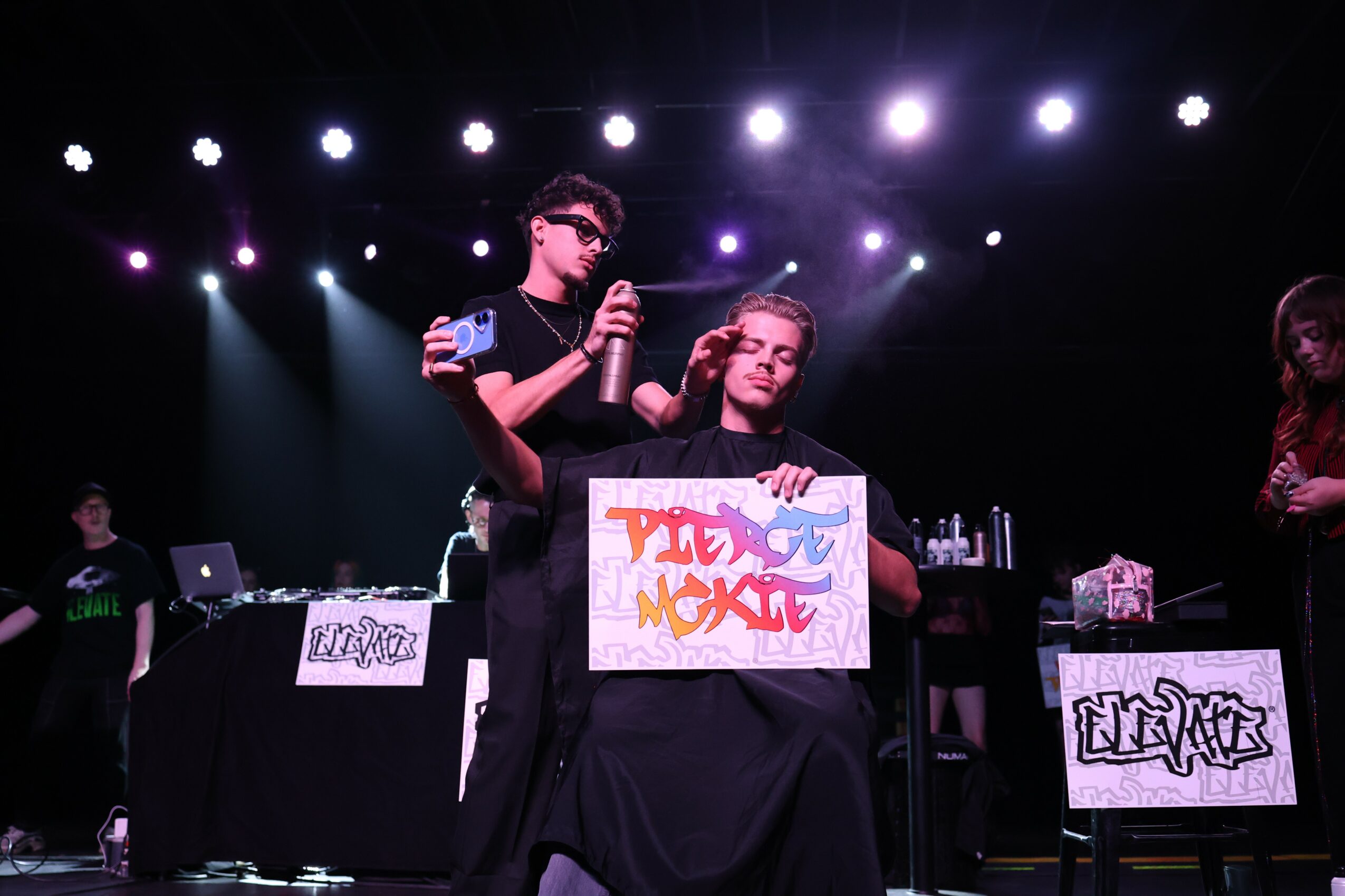 Person sitting on a chair holding a colorful sign, with a person standing behind them holding a microphone, stage lights overhead.