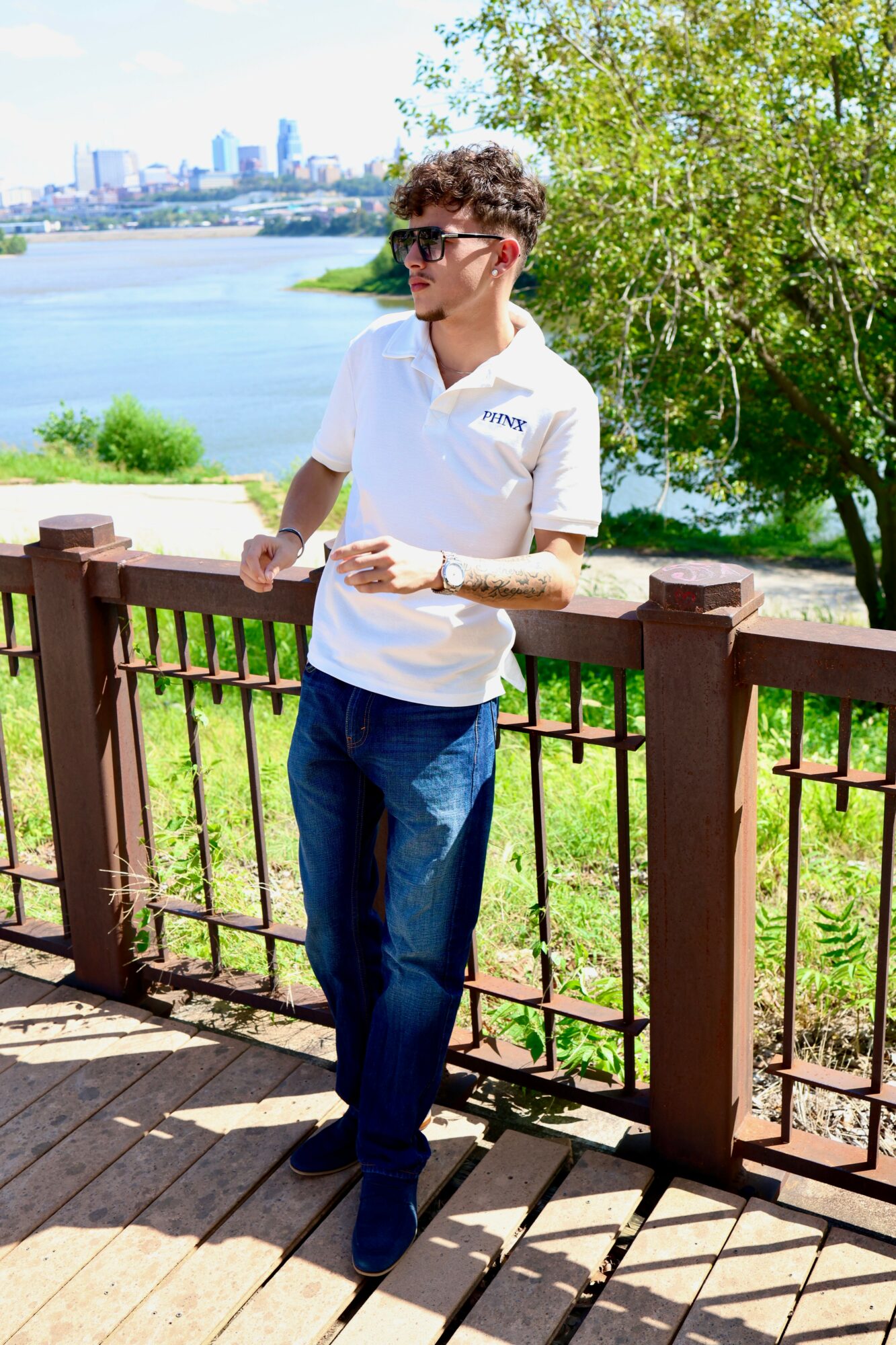 Young man with curly hair and sunglasses leaning on a wooden railing near a river, with trees and city skyline in background.