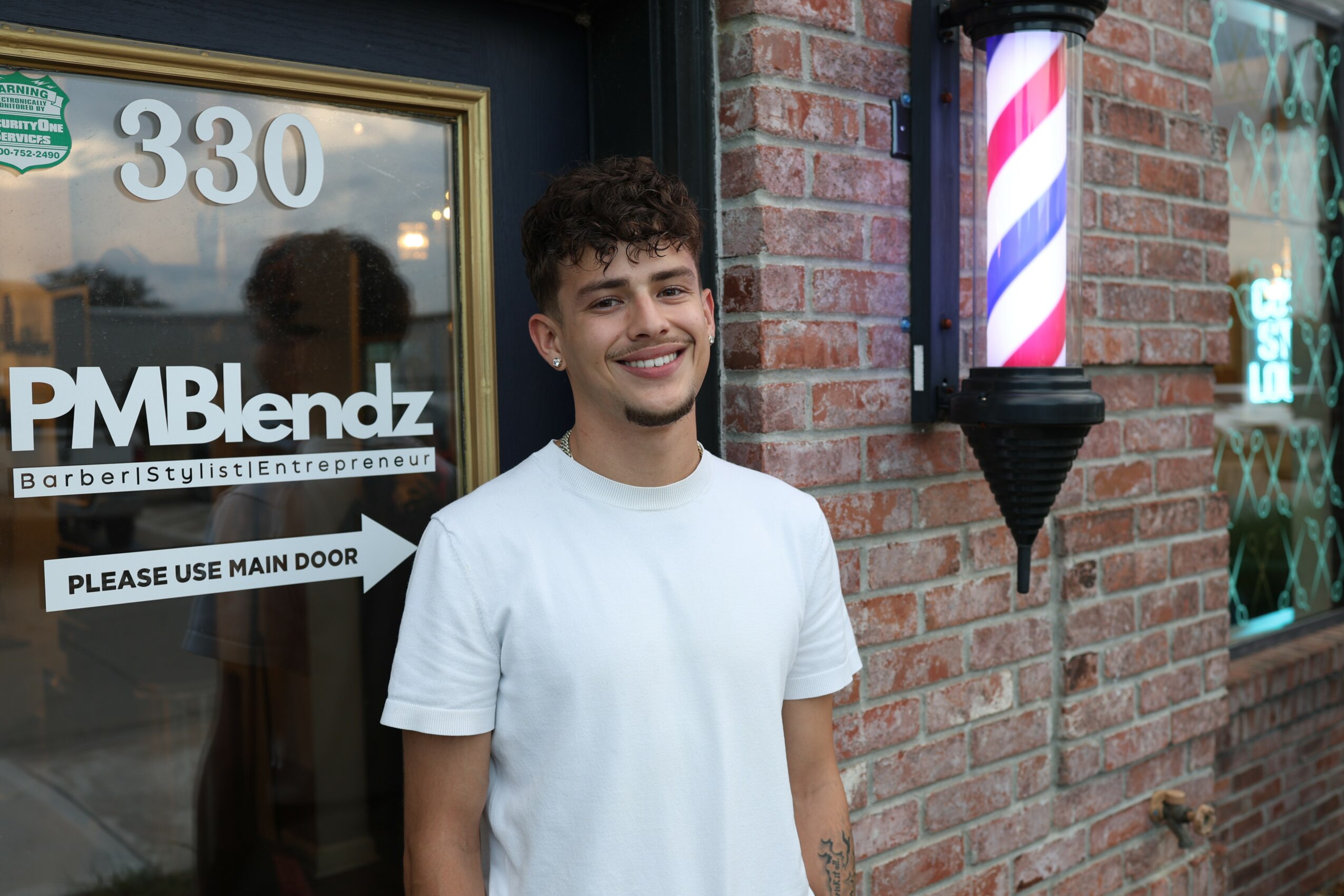 Young man with curly hair and a white t-shirt standing outside a building with a brick wall and barber pole, smiling.