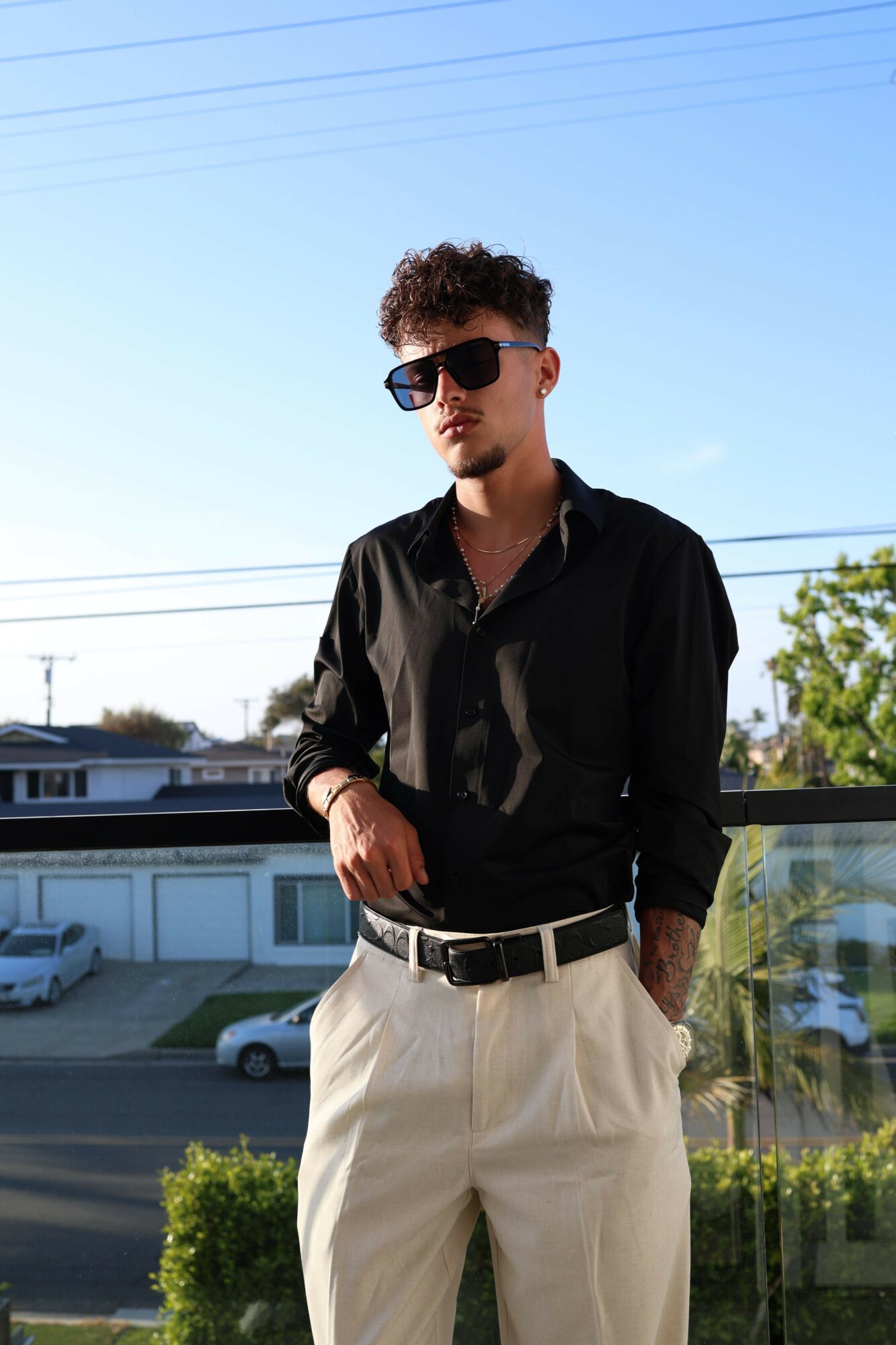 Young man with sunglasses and curly hair stands outdoors, wearing a black shirt and beige pants, with a street and trees in background.