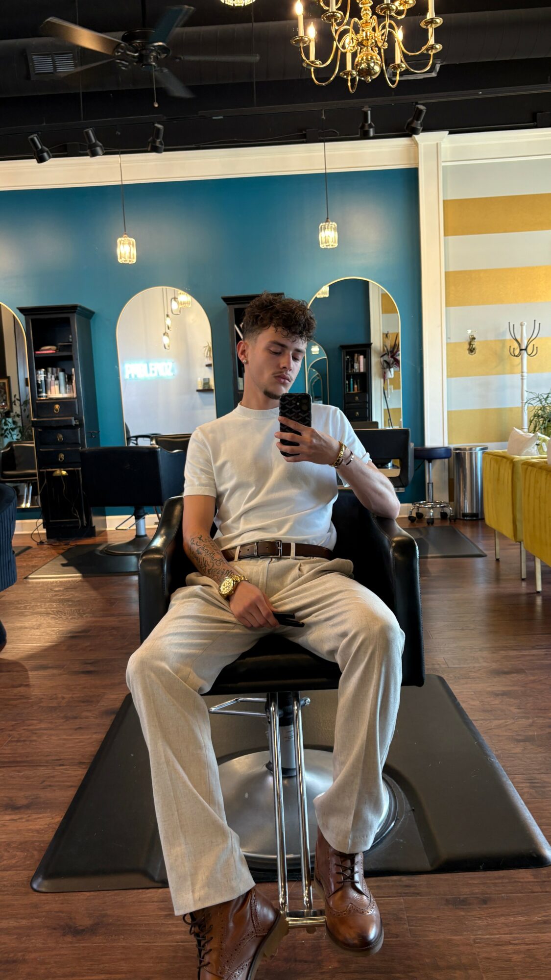 Young man sitting in a salon chair taking a selfie, with mirrors and colorful decor in the background.