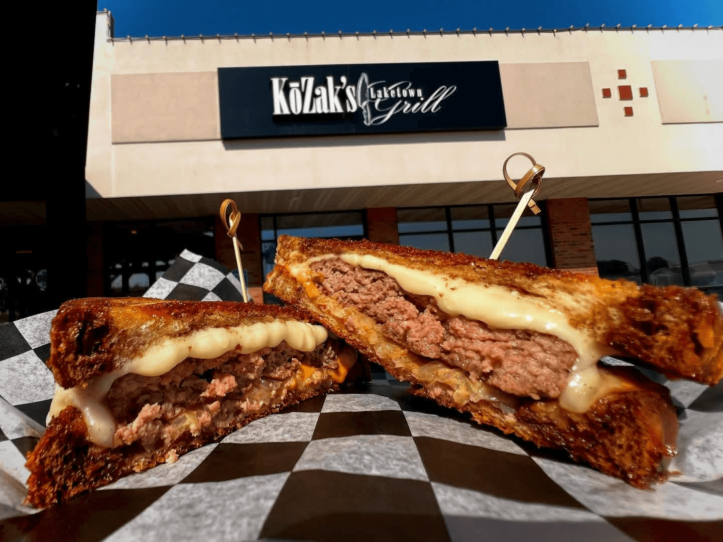 Close-up of a cheeseburger cut in half, showing melted cheese and beef patty, on checkered paper outside restaurant.